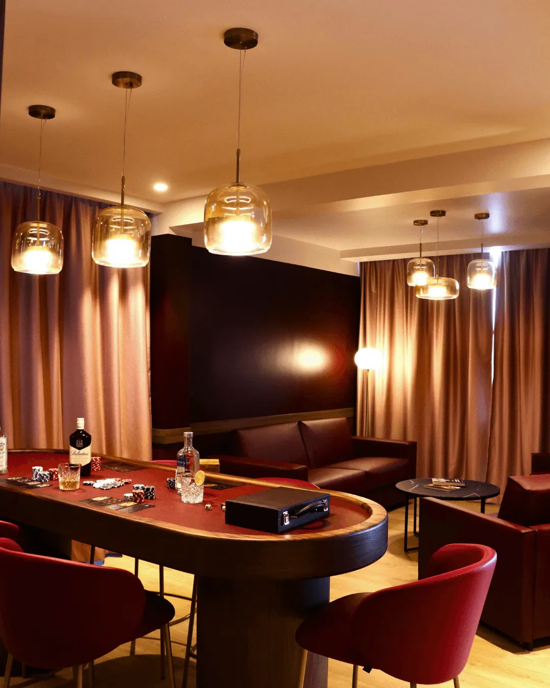 Salle de jeux avec table de poker, chaises rouges, luminaires suspendus et canapés en cuir marron