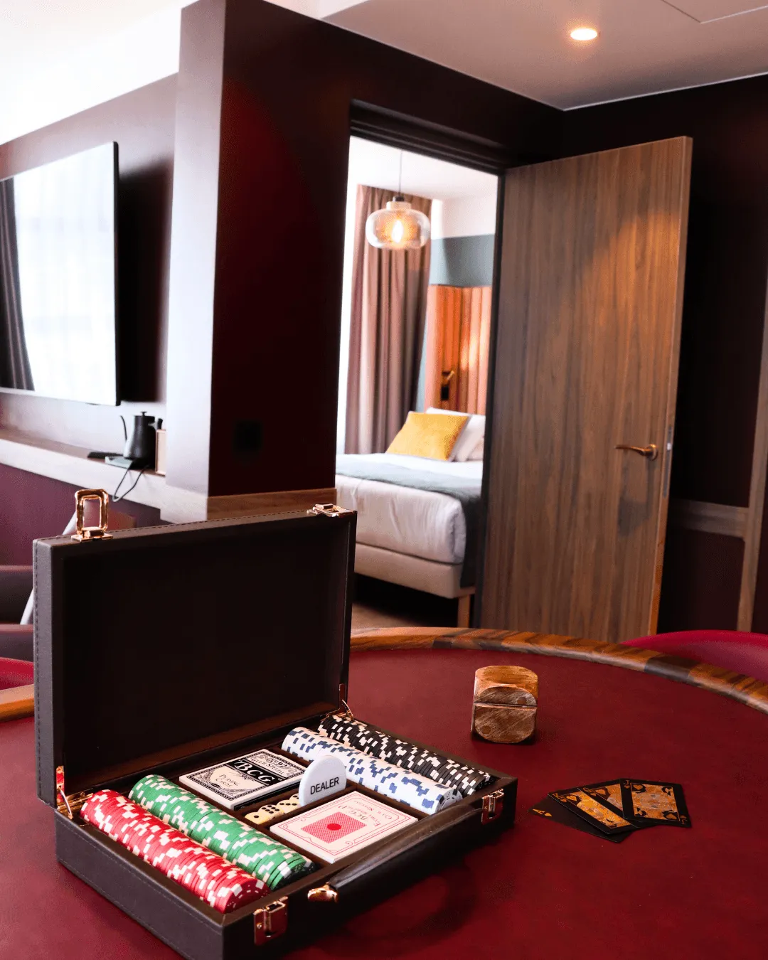 Une table de jeu avec un coffret ouvert contenant des jetons, des cartes et un jeton "DEALER", dans une chambre d'hôtel avec un lit visible à l'arrière-plan