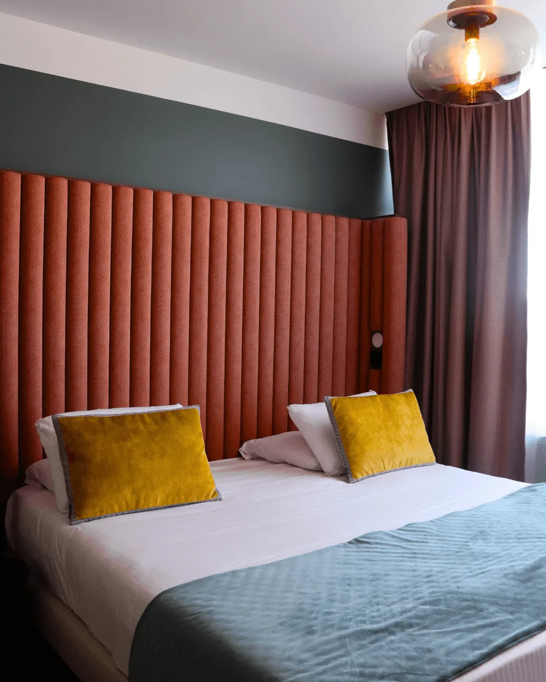 Lit avec une tête de lit rouge rayée, des draps blancs et une couverture grise, deux coussins jaunes et une lampe sphérique allumée au plafond