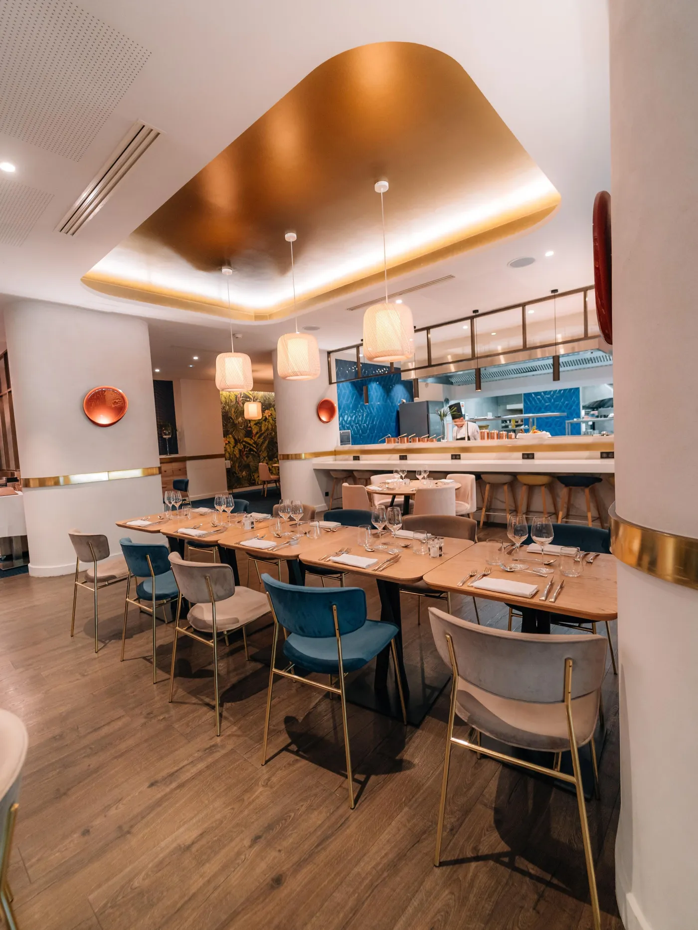Restaurant moderne avec tables dressées, chaises bleues et beiges, éclairage suspendu et cuisine ouverte en arrière-plan