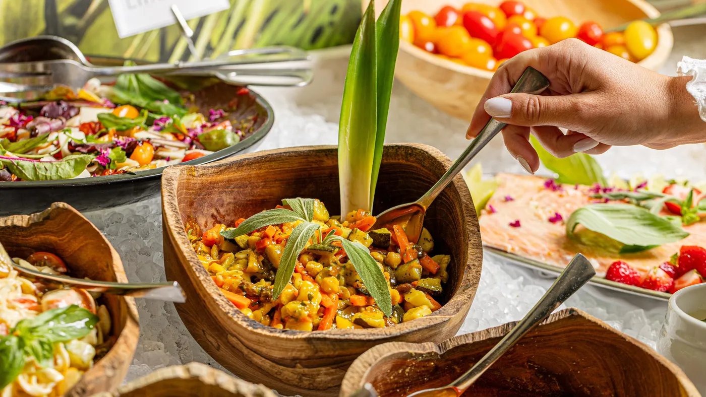 Une personne sert un plat de légumes dans un bol en bois sur une table de buffet avec divers autres plats