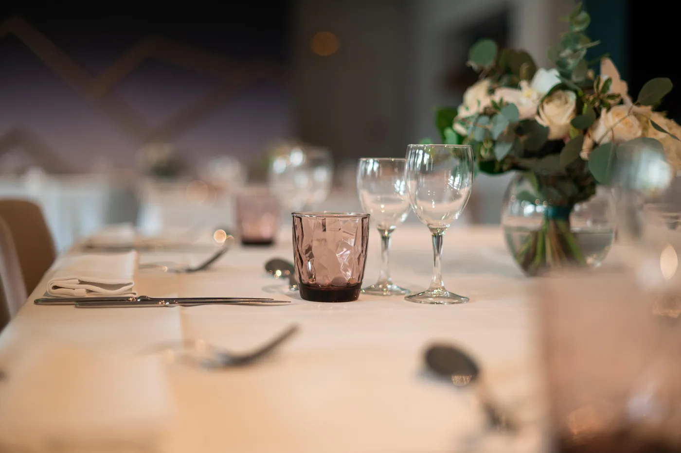 Table dressée avec verres, couverts, nappe blanche et bouquet de fleurs en arrière-plan