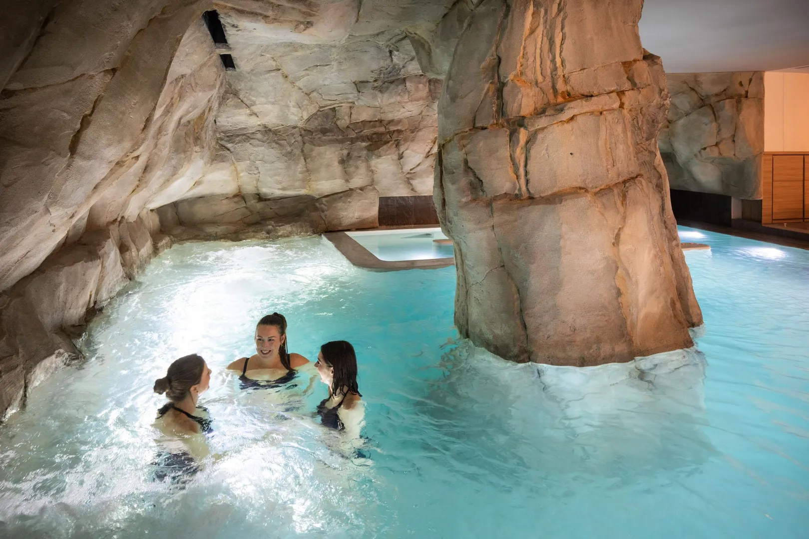 Trois personnes nagent dans une piscine intérieure entourée de rochers artificiels