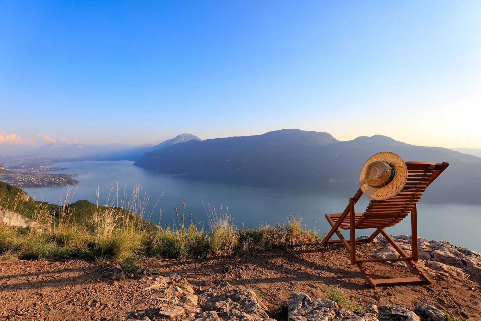 Chaise longue en bois avec un chapeau posé dessus, située sur une colline herbeuse face à un lac et des montagnes au loin