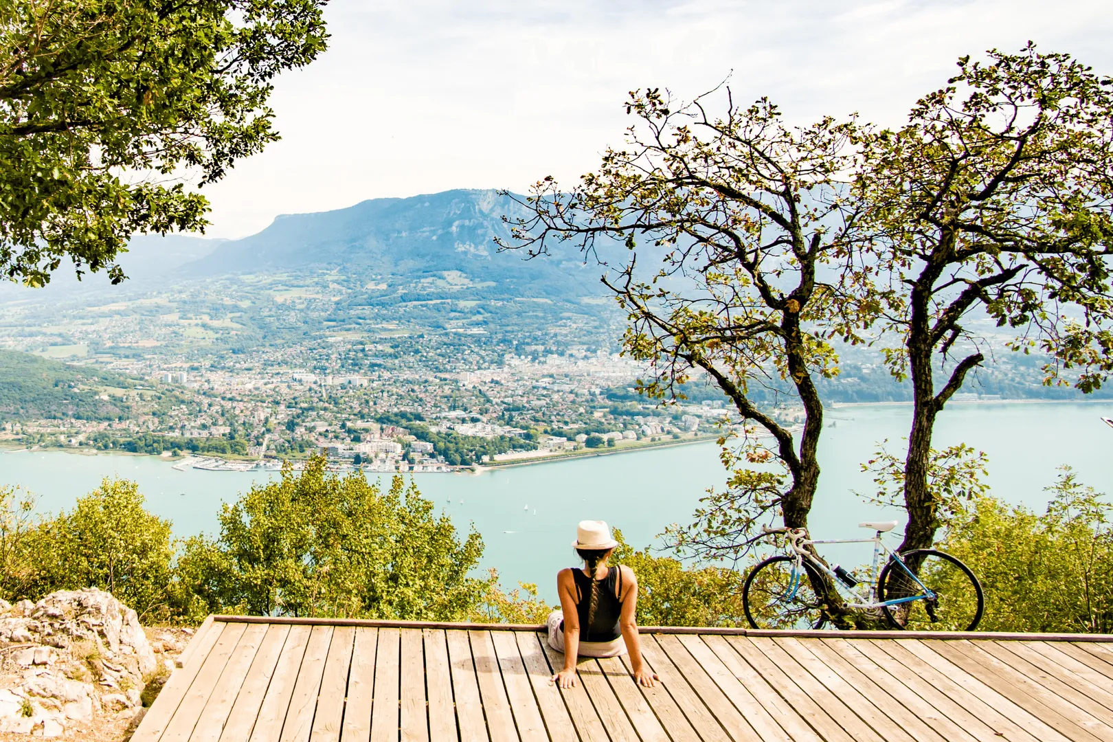 Une personne assise sur une plateforme en bois avec un vélo à côté, face à un lac et une ville entourée de montagnes