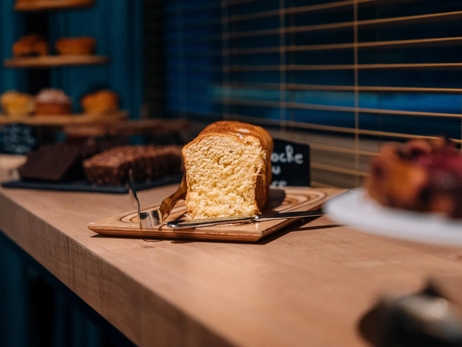 Une tranche de pain posée sur une assiette en bois avec une fourchette et un couteau, devant une vitrine de pâtisserie