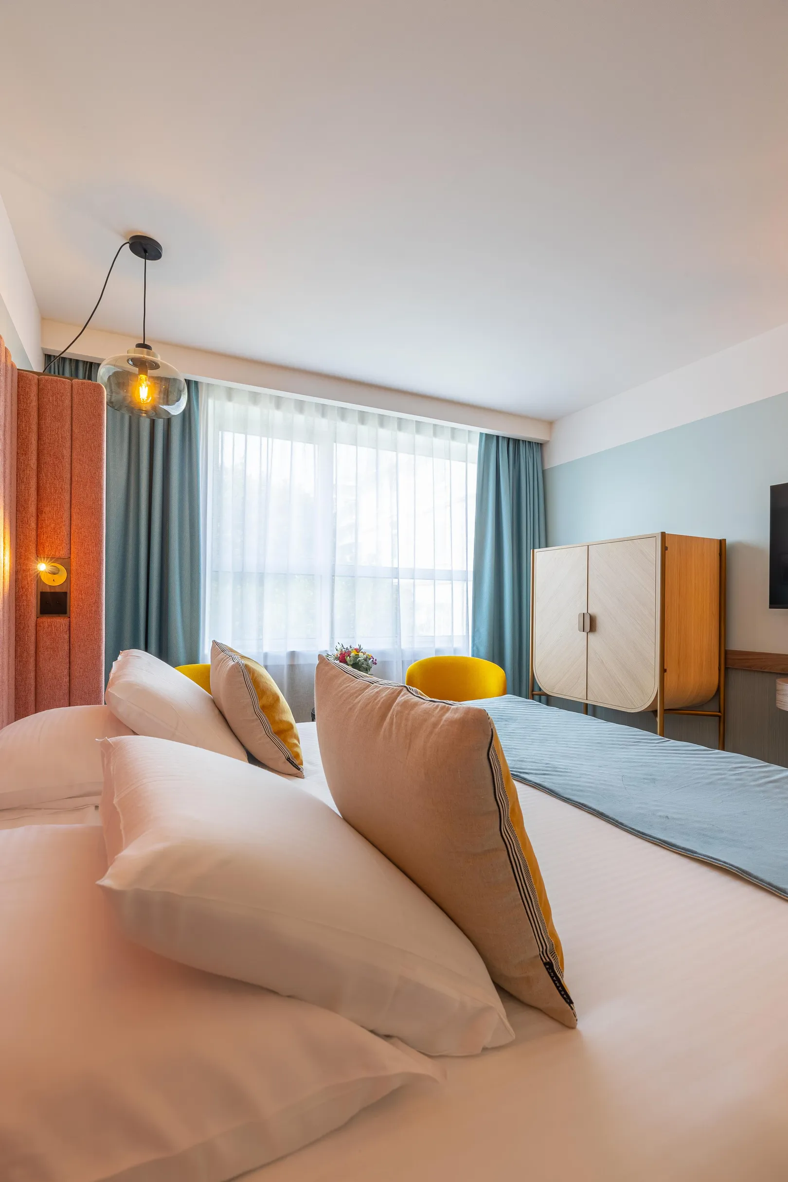 Chambre avec un lit aux couvertures blanches et aux coussins colorés, une fenêtre avec des rideaux, une lampe murale et un meuble en bois
