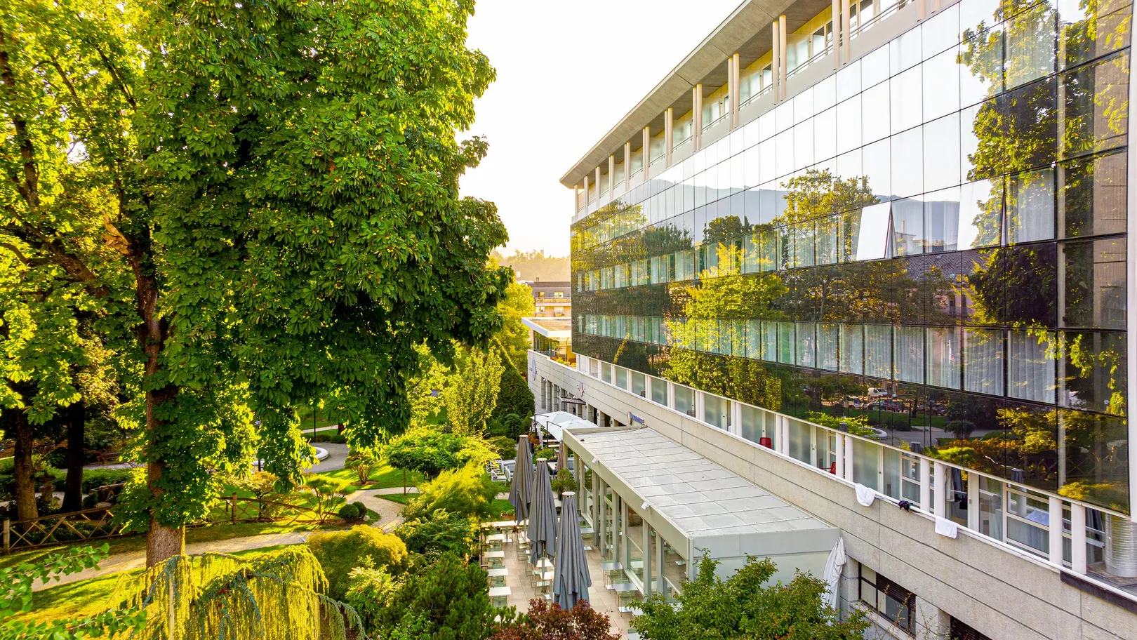 Un bâtiment moderne avec de grandes fenêtres reflétant des arbres verts, adjacent à une terrasse extérieure avec des tables et des chaises