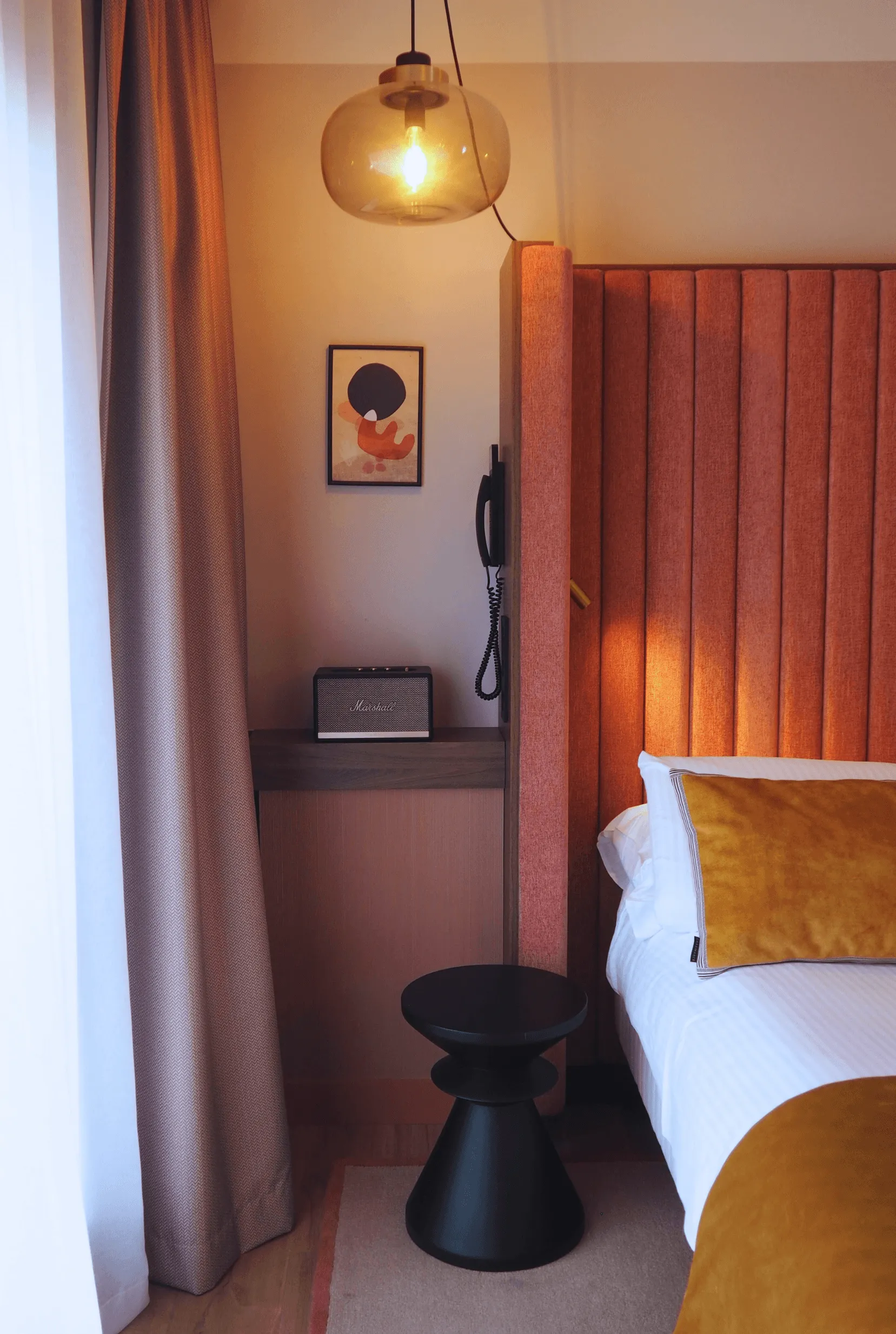 Chambre avec un lit à tête en velours orange, une table de chevet noire, un poste radio et un téléphone sur une étagère, une lampe suspendue et une œuvre d'art au mur