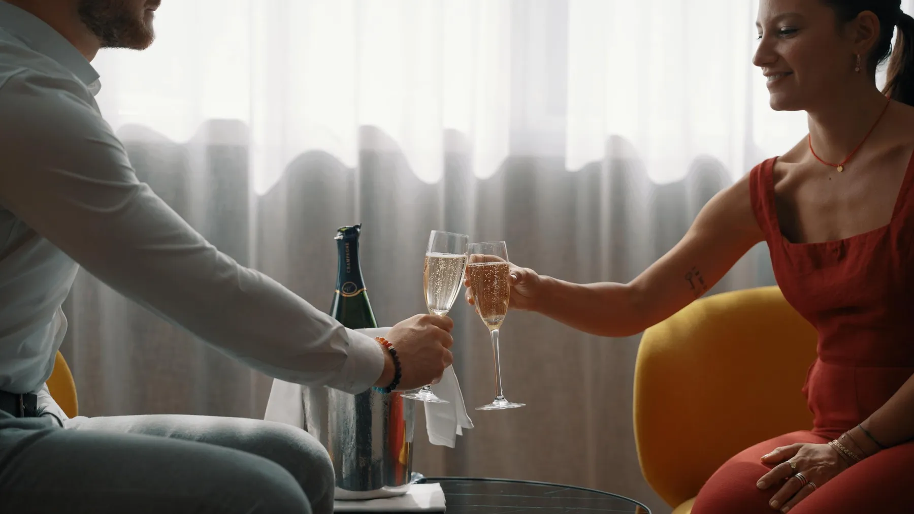 Deux personnes assises sur des chaises, trinquent avec des flûtes à champagne, une bouteille de champagne est posée sur la table entre eux