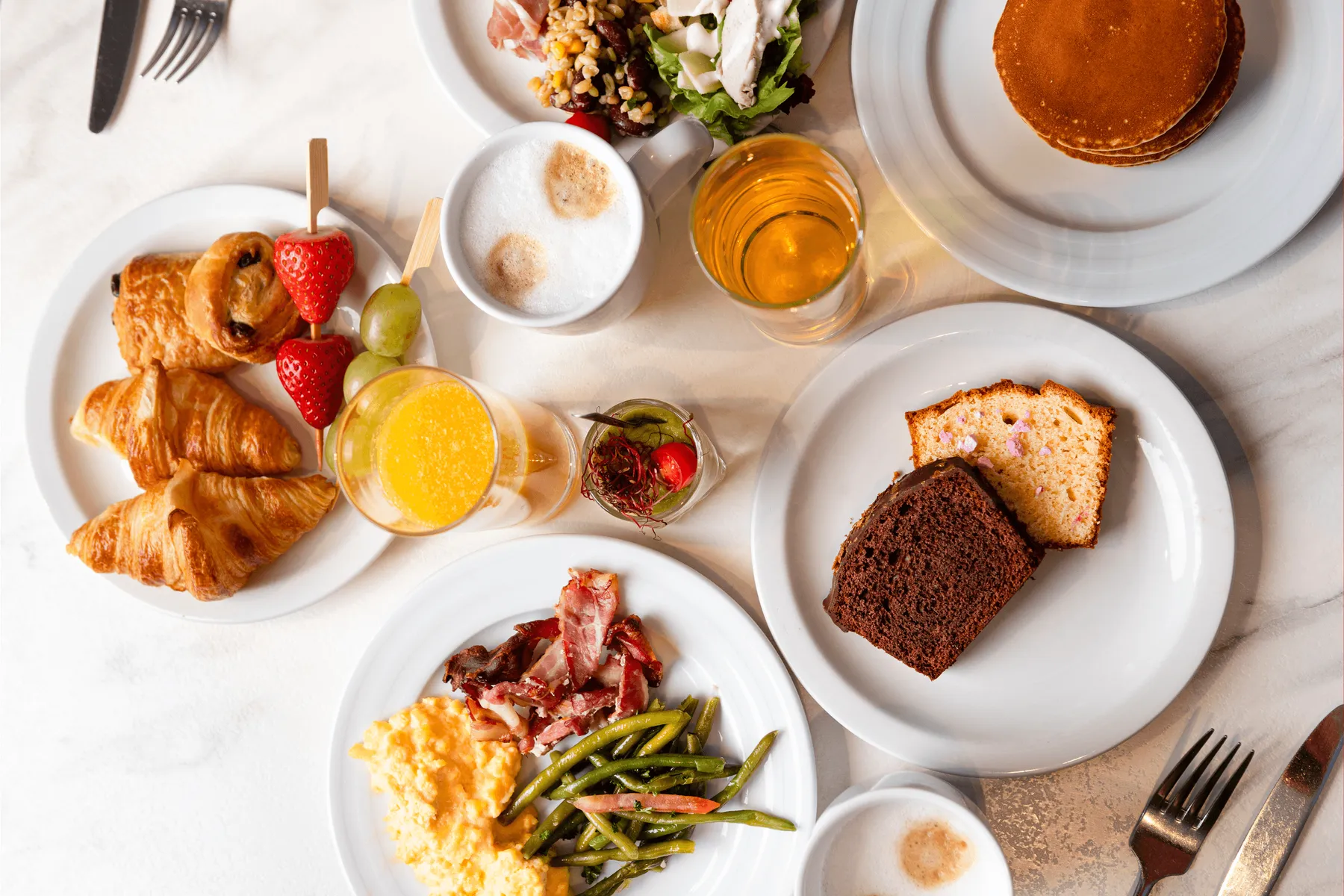 Un assortiment de plats et de boissons disposés sur une table, incluant des croissants, des fruits, des légumes, des œufs, du pain et des boissons chaudes et froides