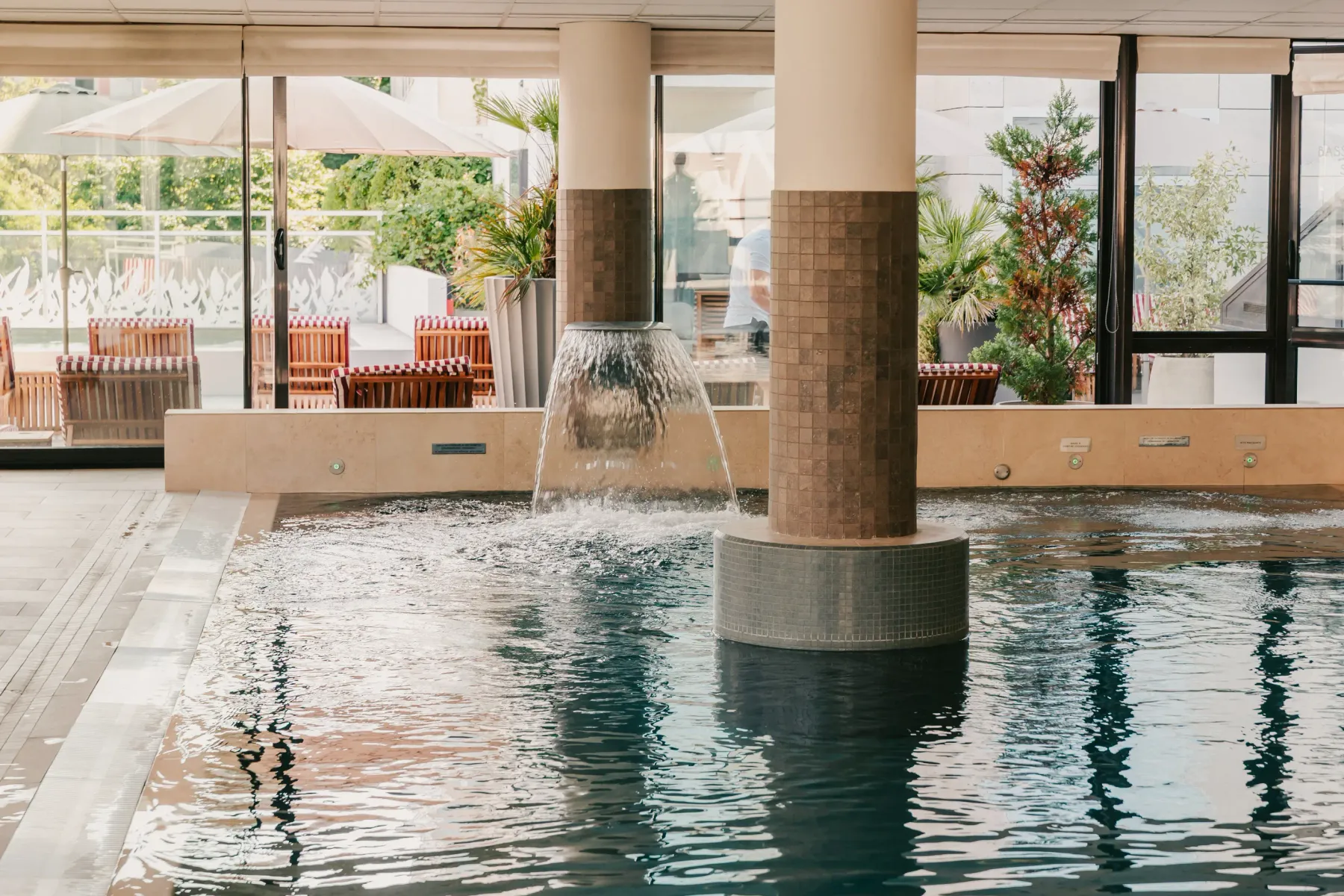 Piscine intérieure avec une fontaine centrale et des tables et chaises en terrasse visibles à travers les grandes baies vitrées