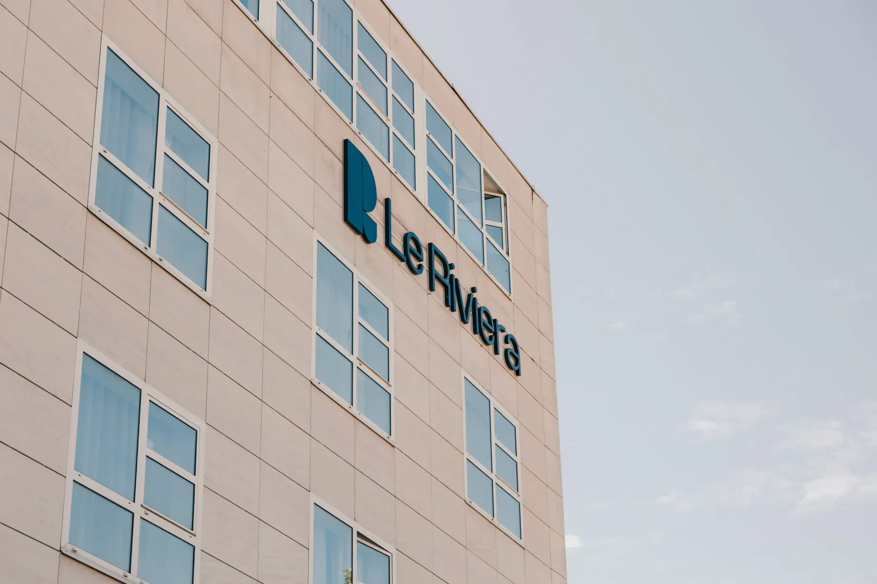 Bâtiment beige avec des fenêtres rectangulaires affichant le logo et le nom "Le Riviera" en bleu