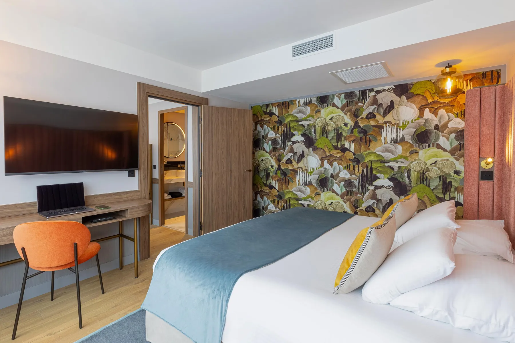 Chambre d'hôtel avec un lit, une chaise orange, un bureau avec un ordinateur portable, une télévision et une porte ouverte sur une salle de bain