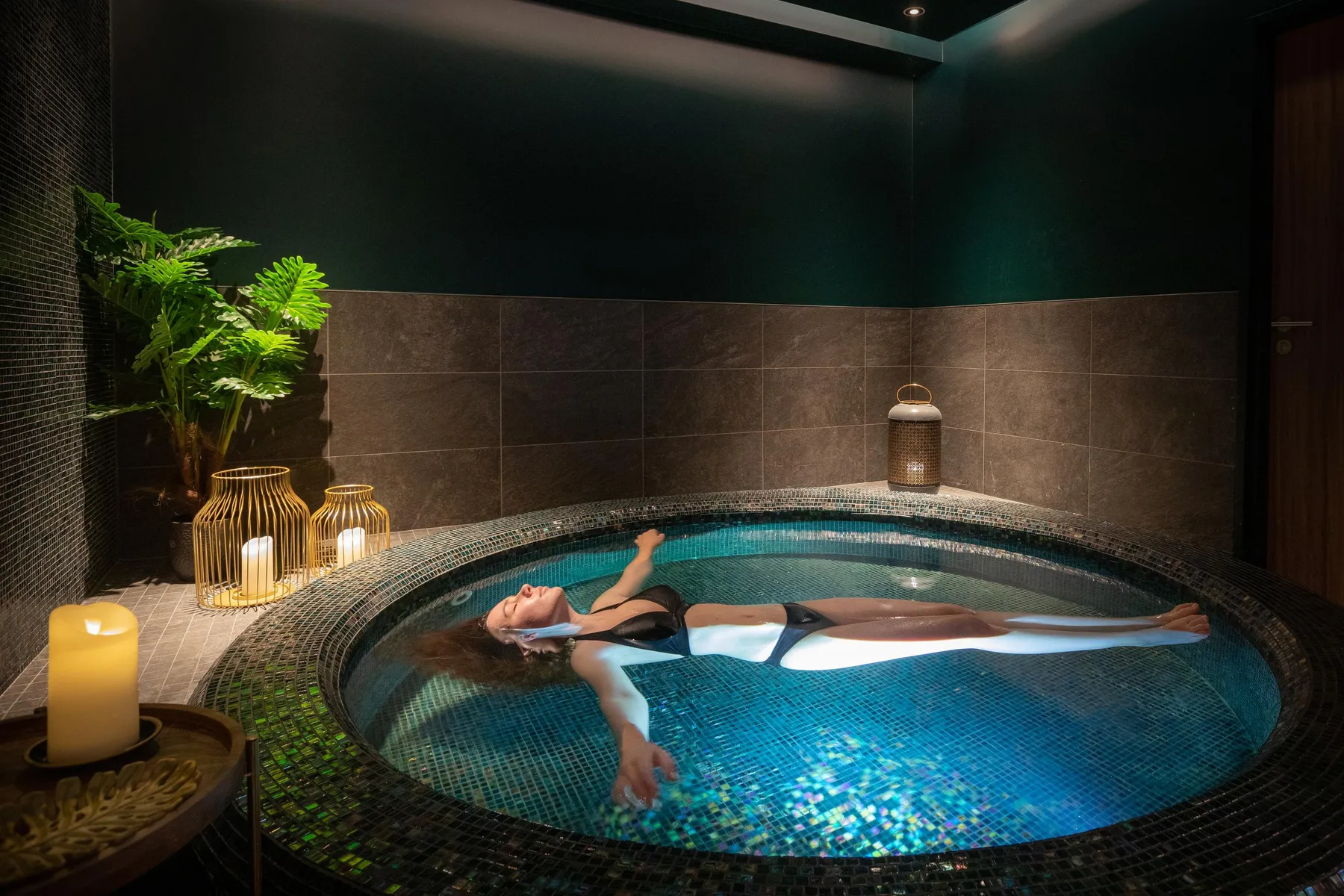 Une personne allongée dans un bain à remous circulaire entouré de bougies et de plantes vertes