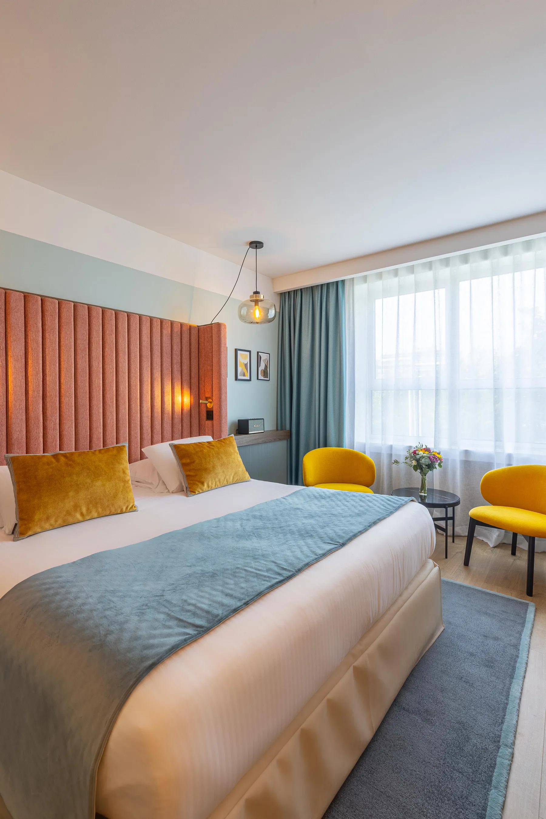 Chambre d'hôtel avec un lit double, deux fauteuils jaunes, une table basse avec un vase de fleurs, des rideaux et une grande fenêtre