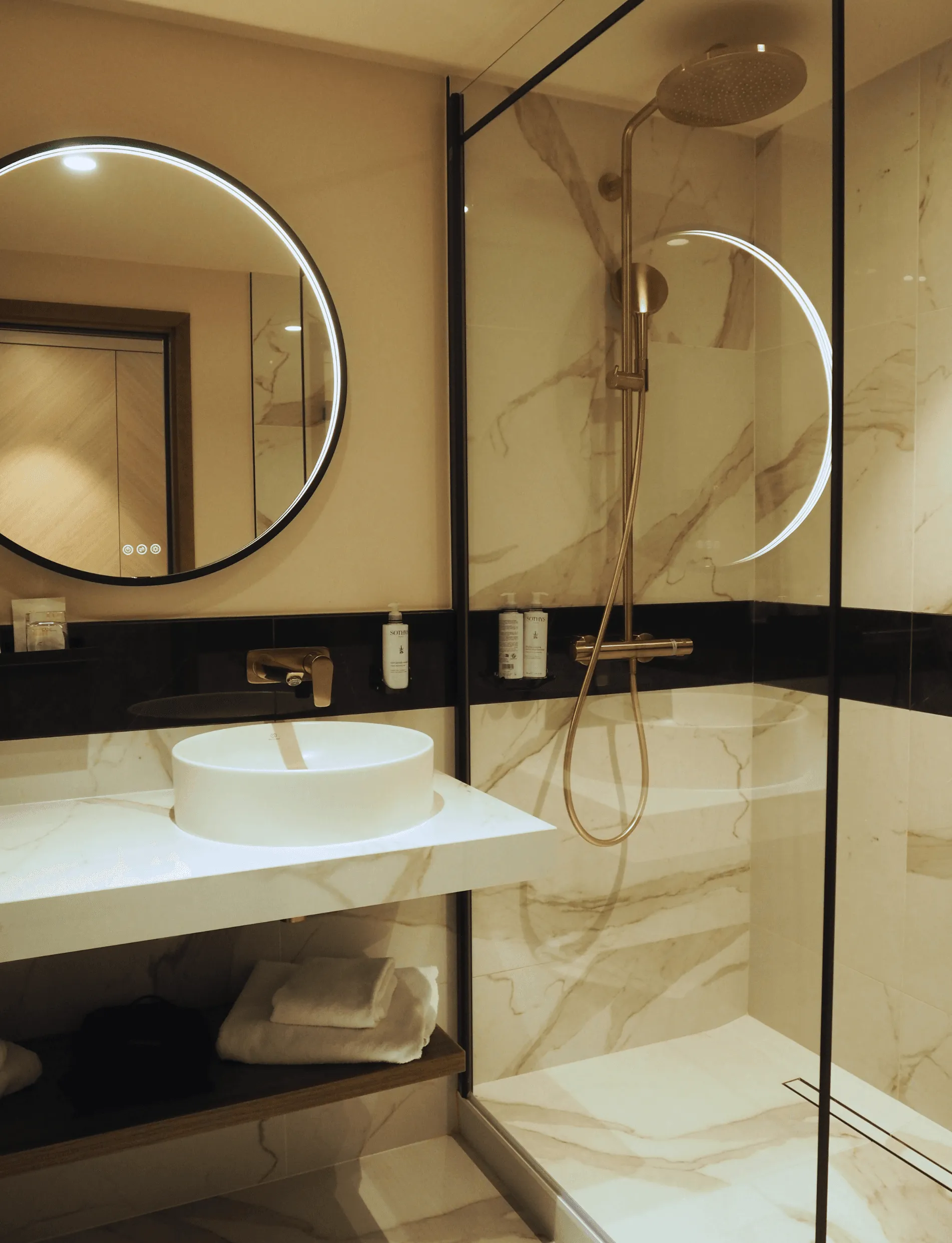 Salle de bain moderne avec lavabo blanc, miroir rond éclairé et douche à effet pluie derrière une paroi vitrée