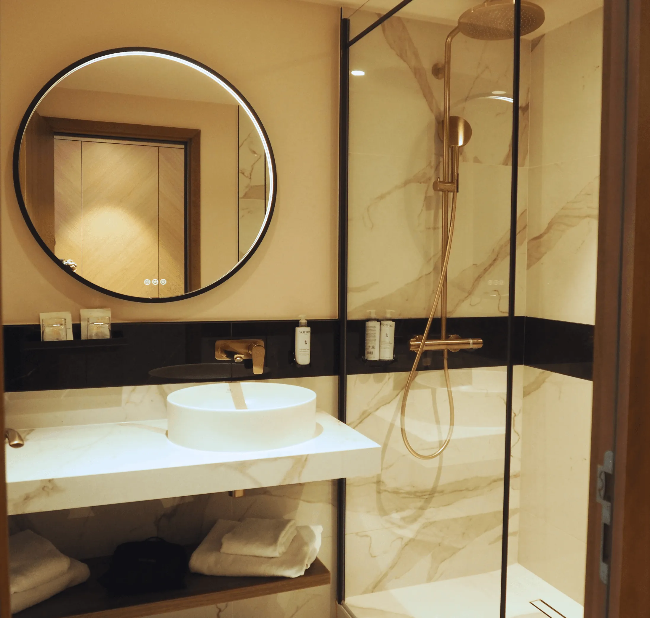 Salle de bain avec lavabo blanc, miroir rond éclairé et douche à paroi en verre