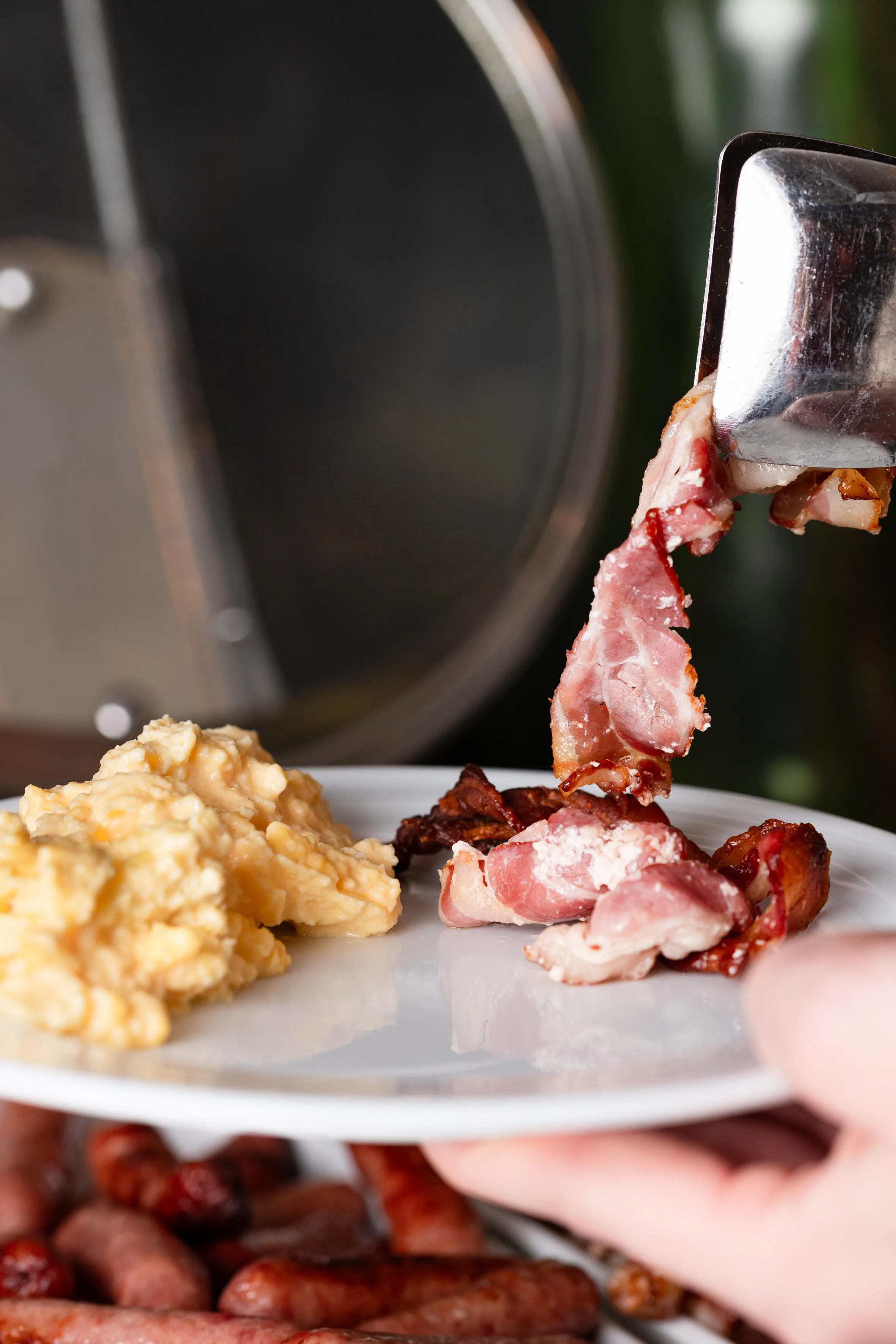 A piece of bacon is picked up by tongs and placed on a plate with scrambled eggs and fried bacon bits.