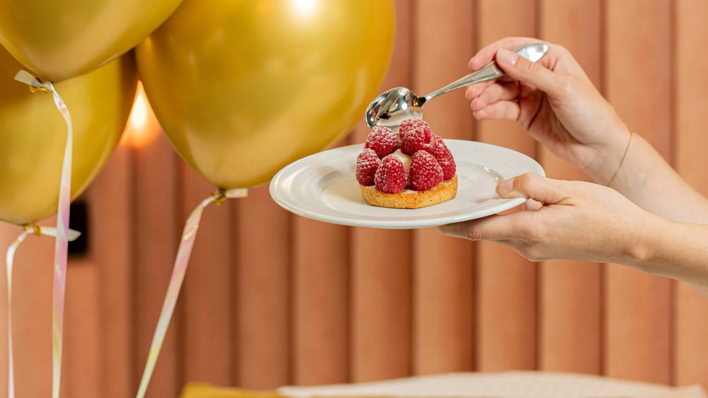 Une personne verse du sirop sur une tartelette garnie de framboises posée sur une assiette blanche, avec des ballons dorés en arrière-plan