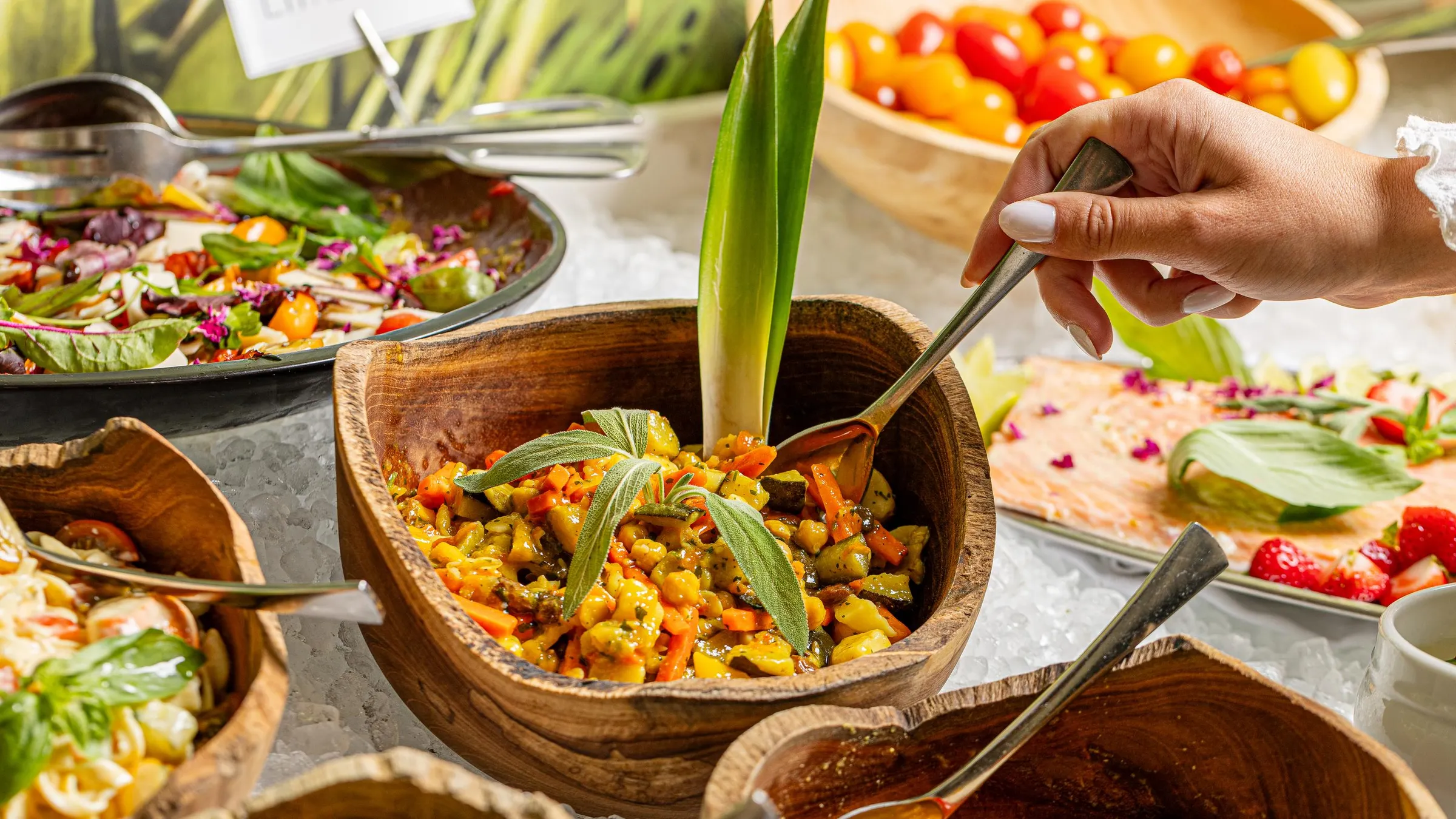 Une personne sert un plat de légumes dans un bol en bois sur une table de buffet avec divers autres plats