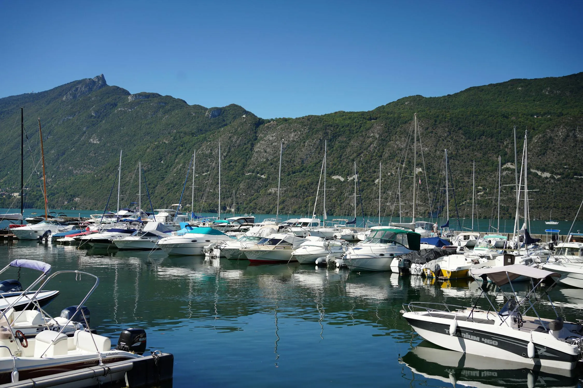 Un port de plaisance avec plusieurs bateaux amarrés, entouré de montagnes verdoyantes sous un ciel bleu clair