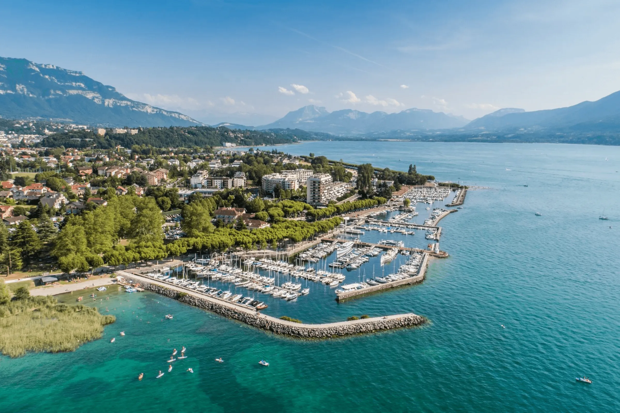 Village au bord d'un lac avec marina, montagnes en arrière-plan et personnes en paddle dans l'eau