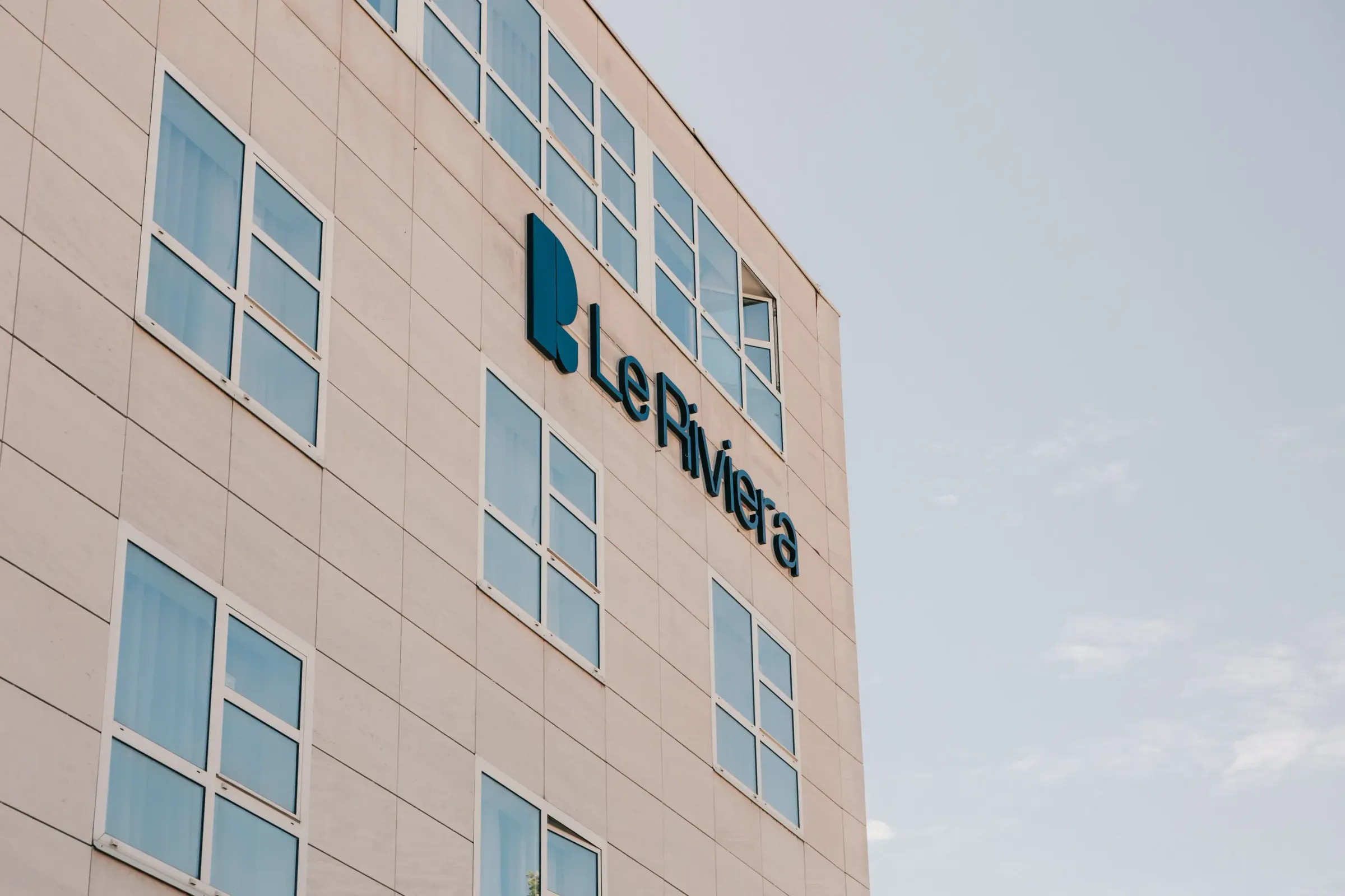 Bâtiment beige avec des fenêtres rectangulaires affichant le logo et le nom "Le Riviera" en bleu