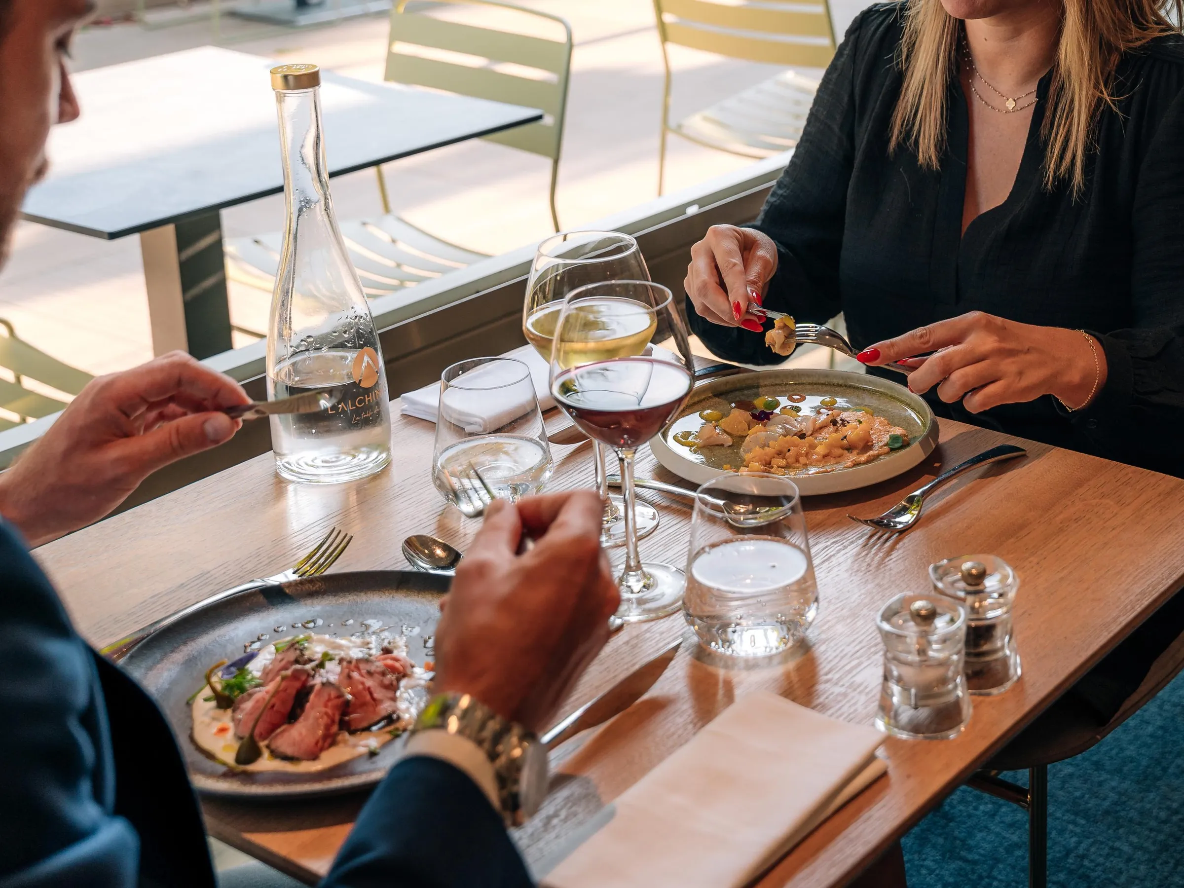 Trois personnes assises à une table en train de manger des plats et de boire du vin, avec des verres, des couverts et des bouteilles sur la table