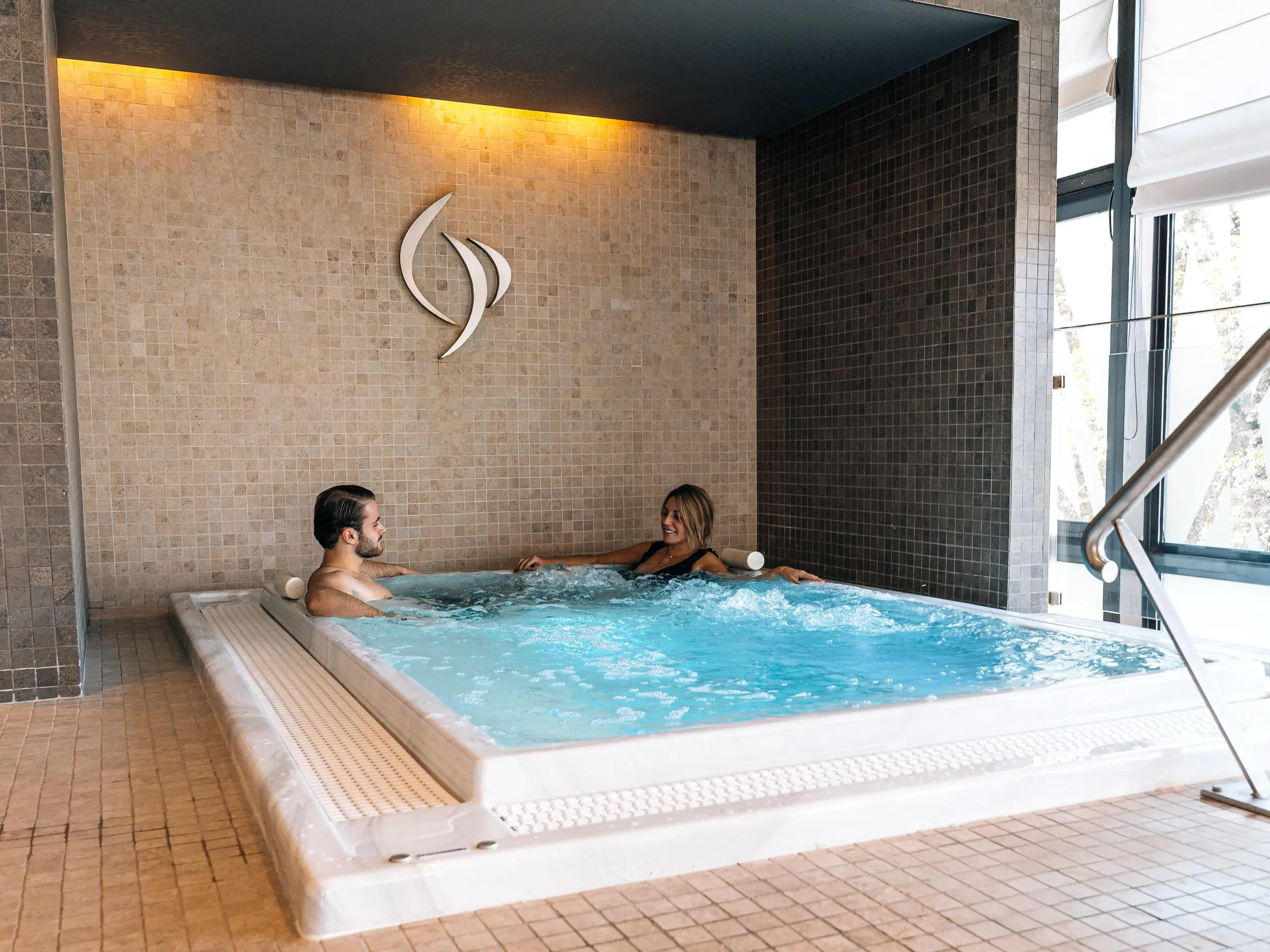 Deux personnes sont assises dans un jacuzzi, entourées de carrelage, avec une grande fenêtre à proximité