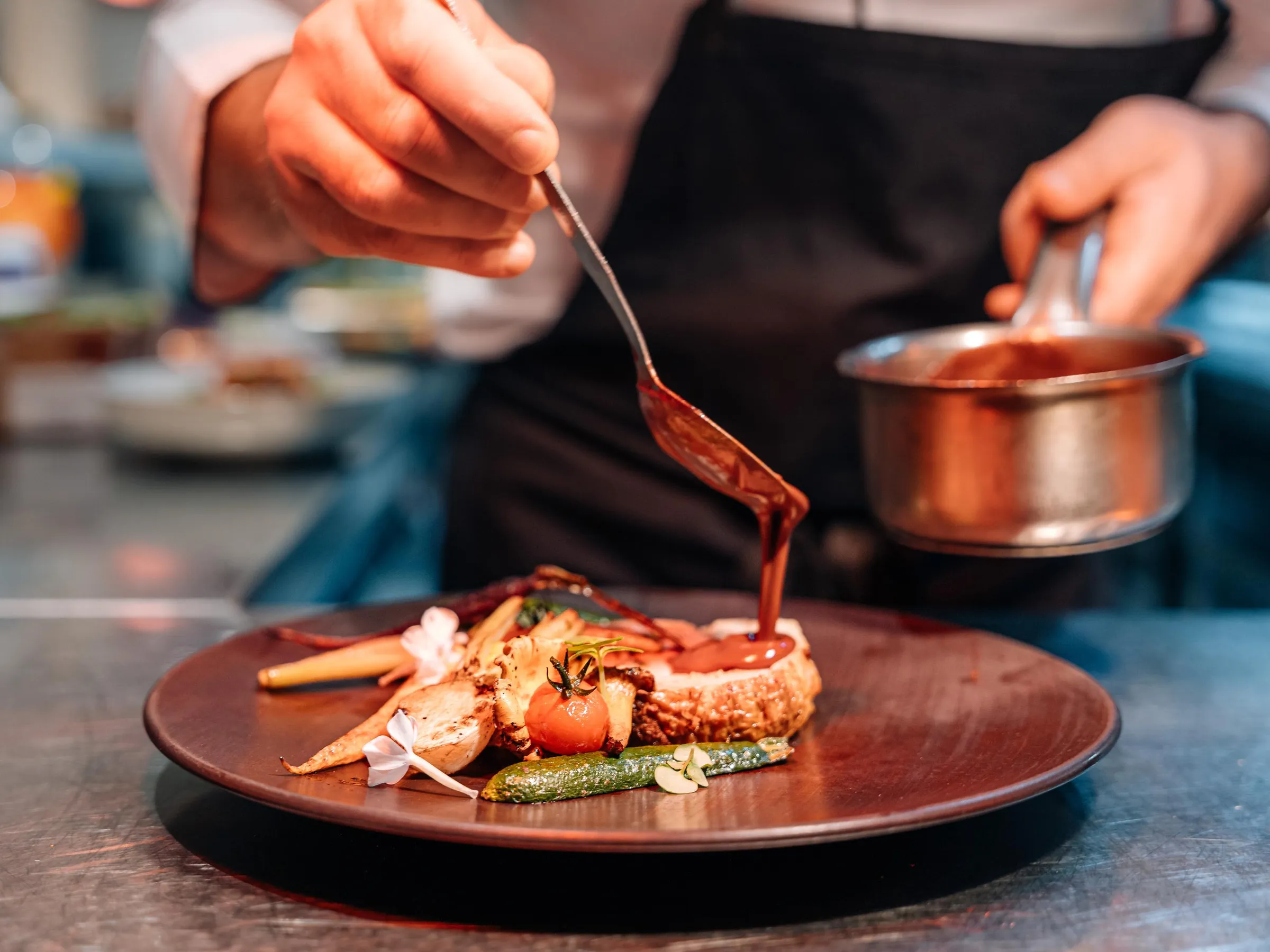 Un chef de cuisine verse une sauce sur un plat composé de viande, légumes et garnitures sur une assiette ronde