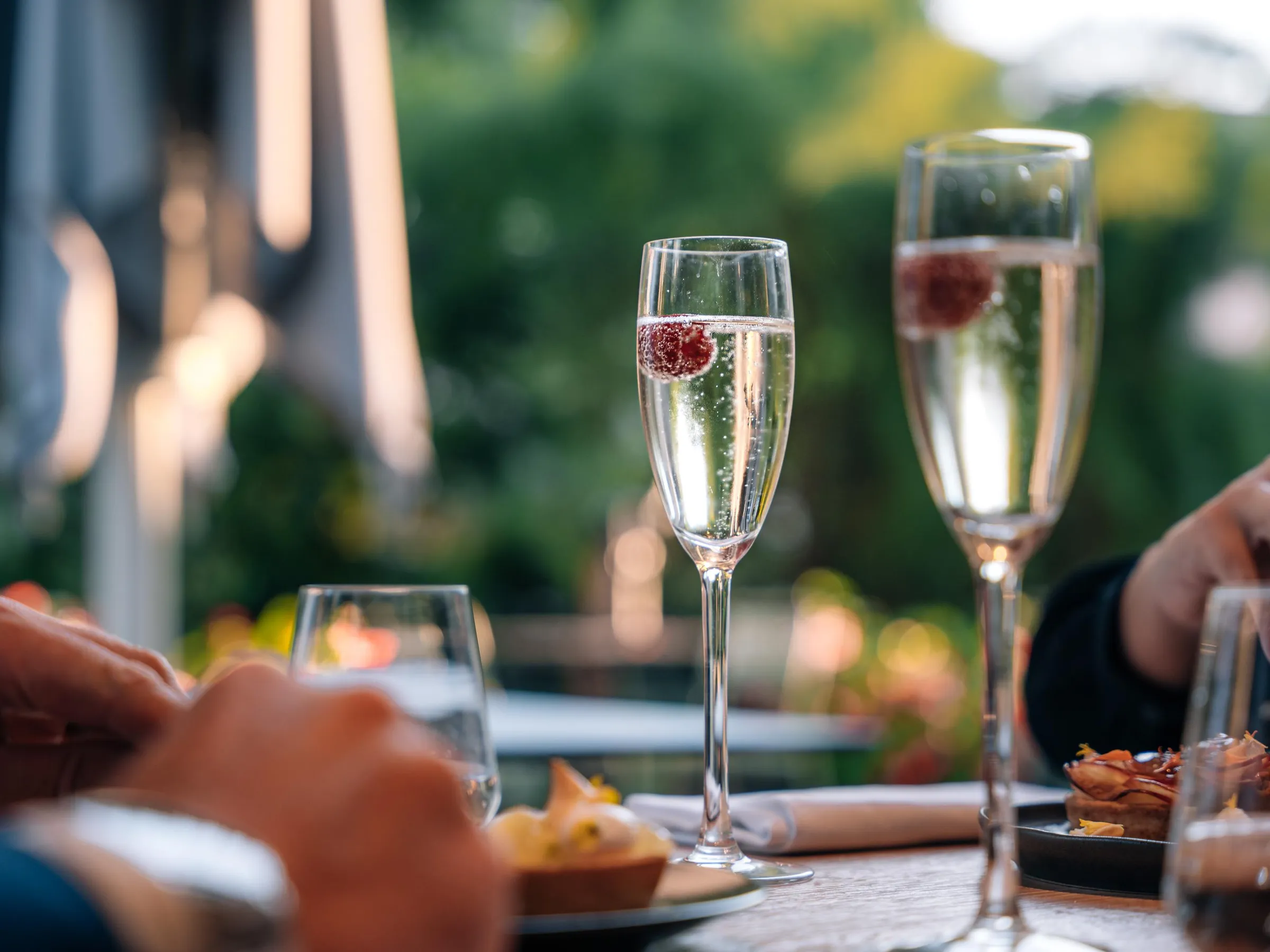 Deux flûtes de champagne avec des fruits rouges à l'intérieur posées sur une table en bois, avec des mains et des verres à vin en arrière-plan