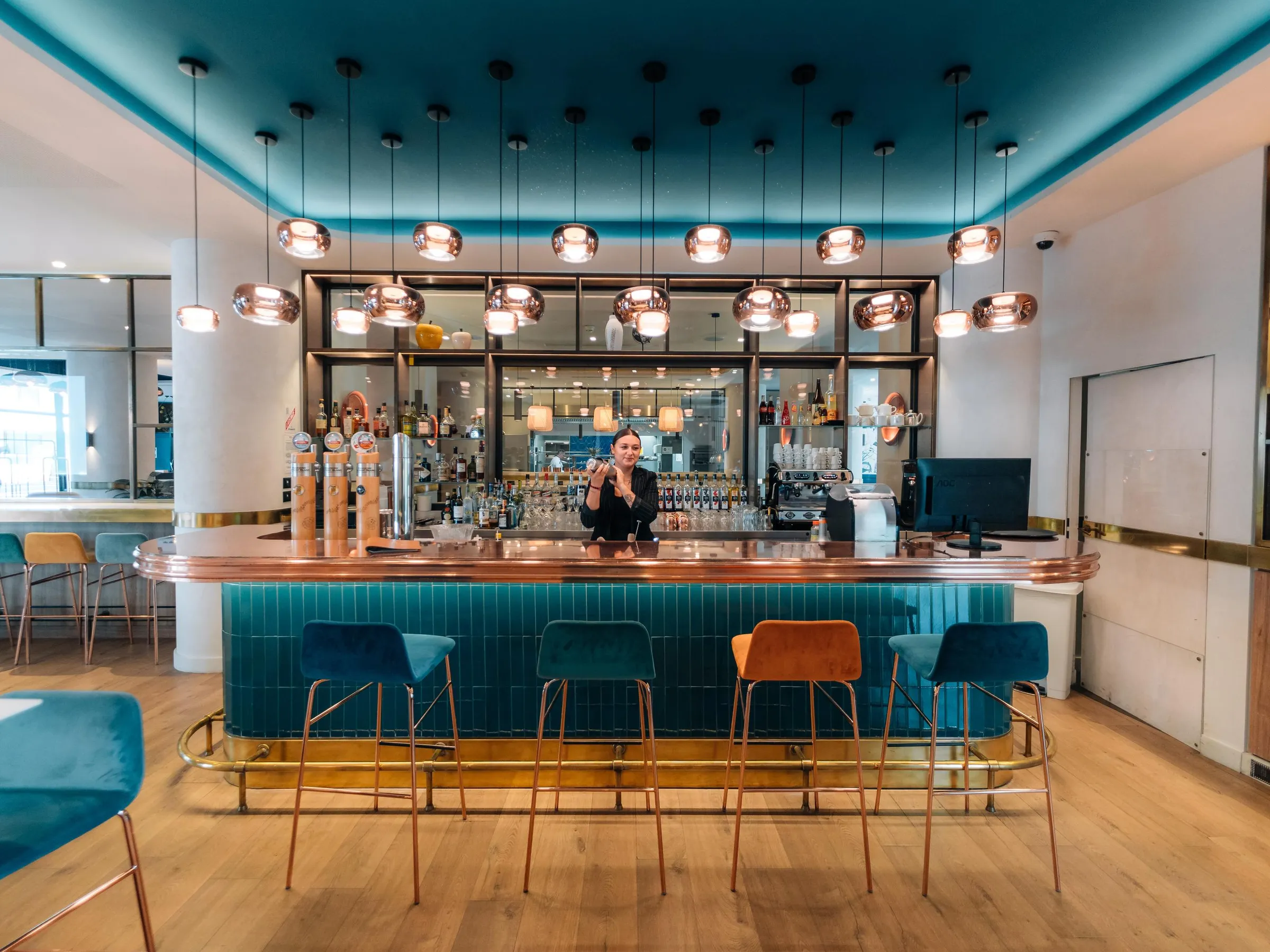 Un bar avec un comptoir en cuivre et des tabourets, une personne derrière le comptoir, des lumières suspendues et des étagères remplies de bouteilles