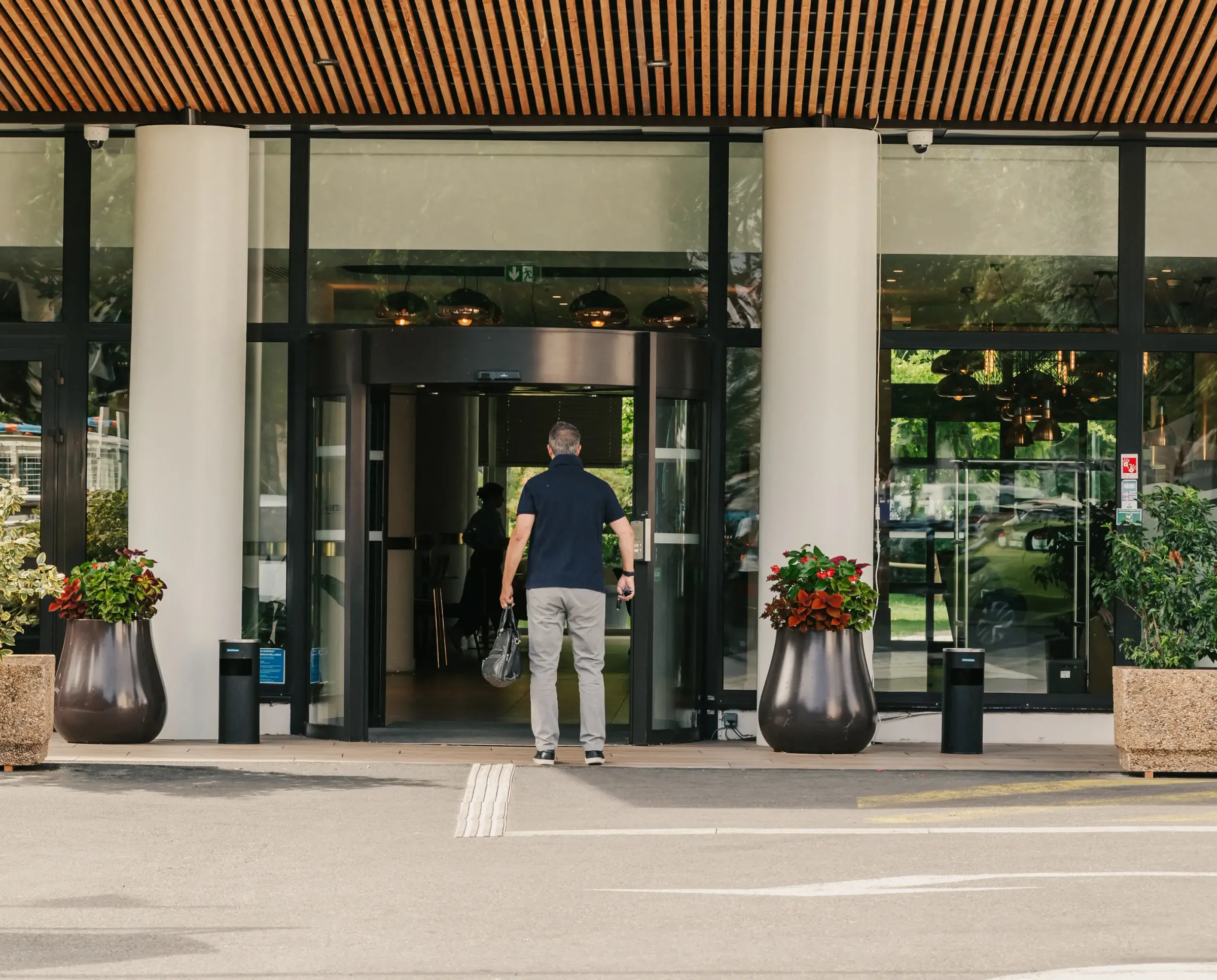 Un homme se tient devant l'entrée d'un bâtiment moderne avec des portes vitrées, des plantes en pot et un trottoir