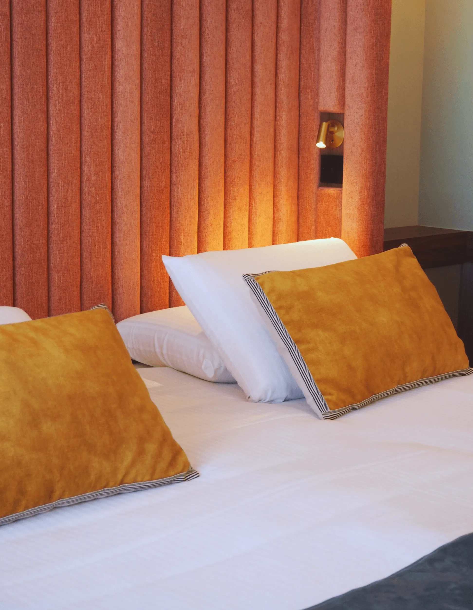 Lit avec des draps blancs et des coussins orange et blancs, un mur avec des rideaux orange et une lampe de chevet allumée