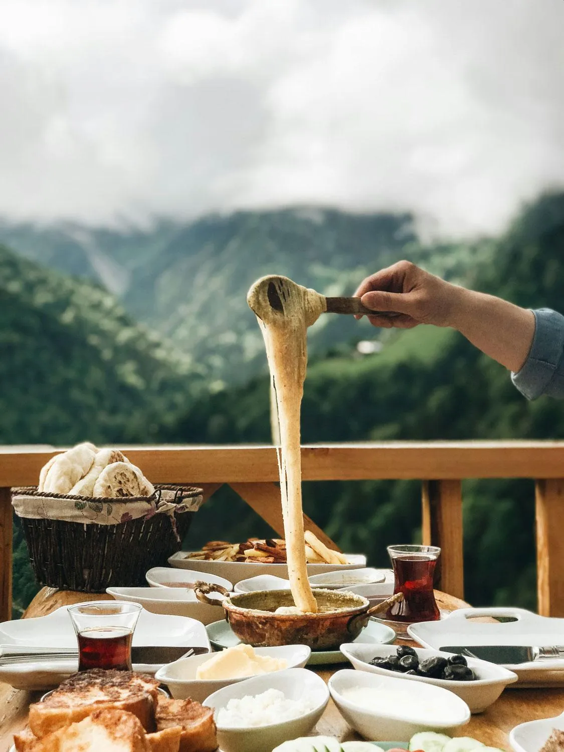 Une main tient une cuillère en bois au-dessus d'un plat contenant une substance crémeuse, sur une table garnie de divers aliments et boissons, avec un paysage montagneux en arrière-plan