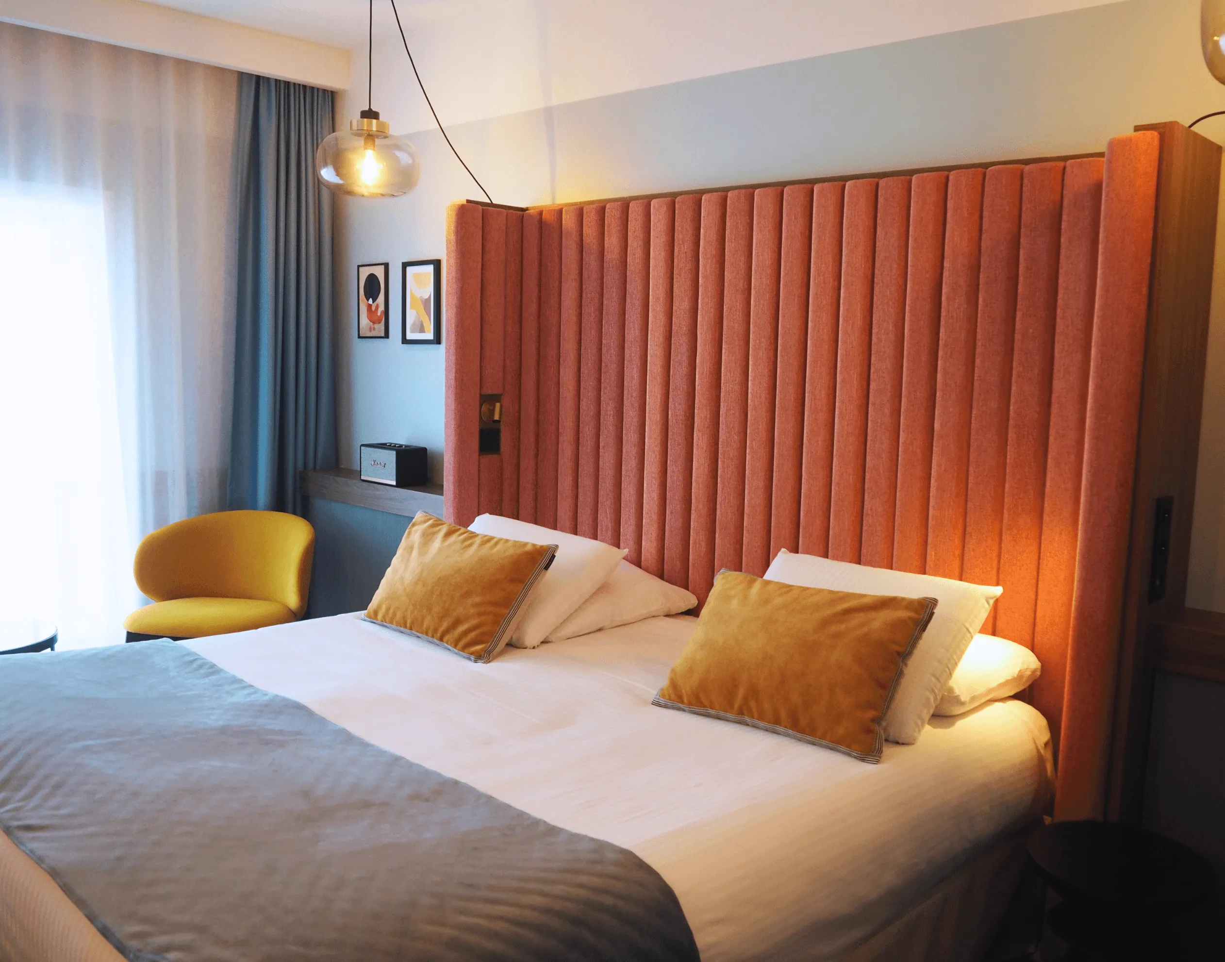 Chambre avec un lit double aux draps blancs et coussins orange, une tête de lit en velours rouge, une chaise jaune et une lampe suspendue