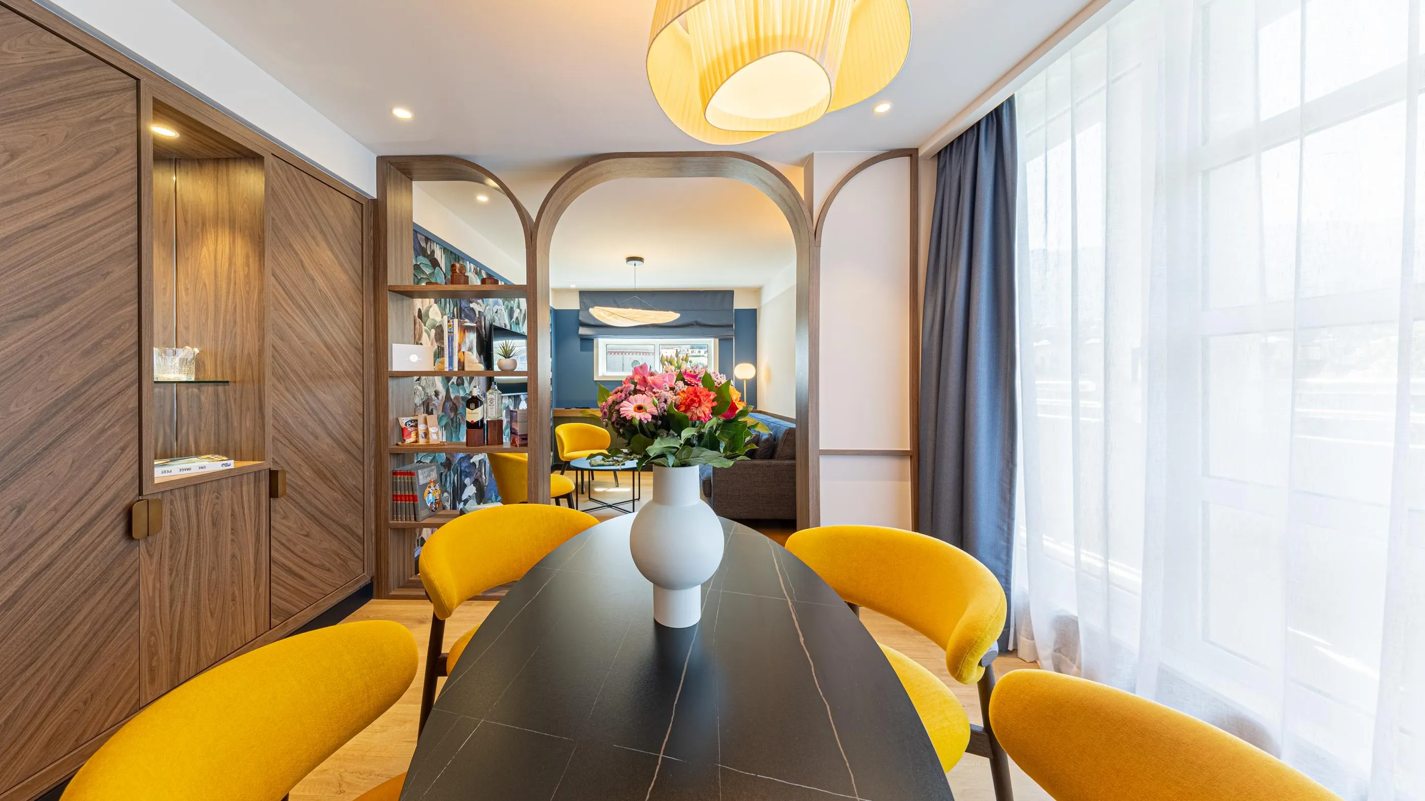 Salle à manger moderne avec table noire, chaises jaunes et vase de fleurs, vue sur un salon bleu à travers des arches