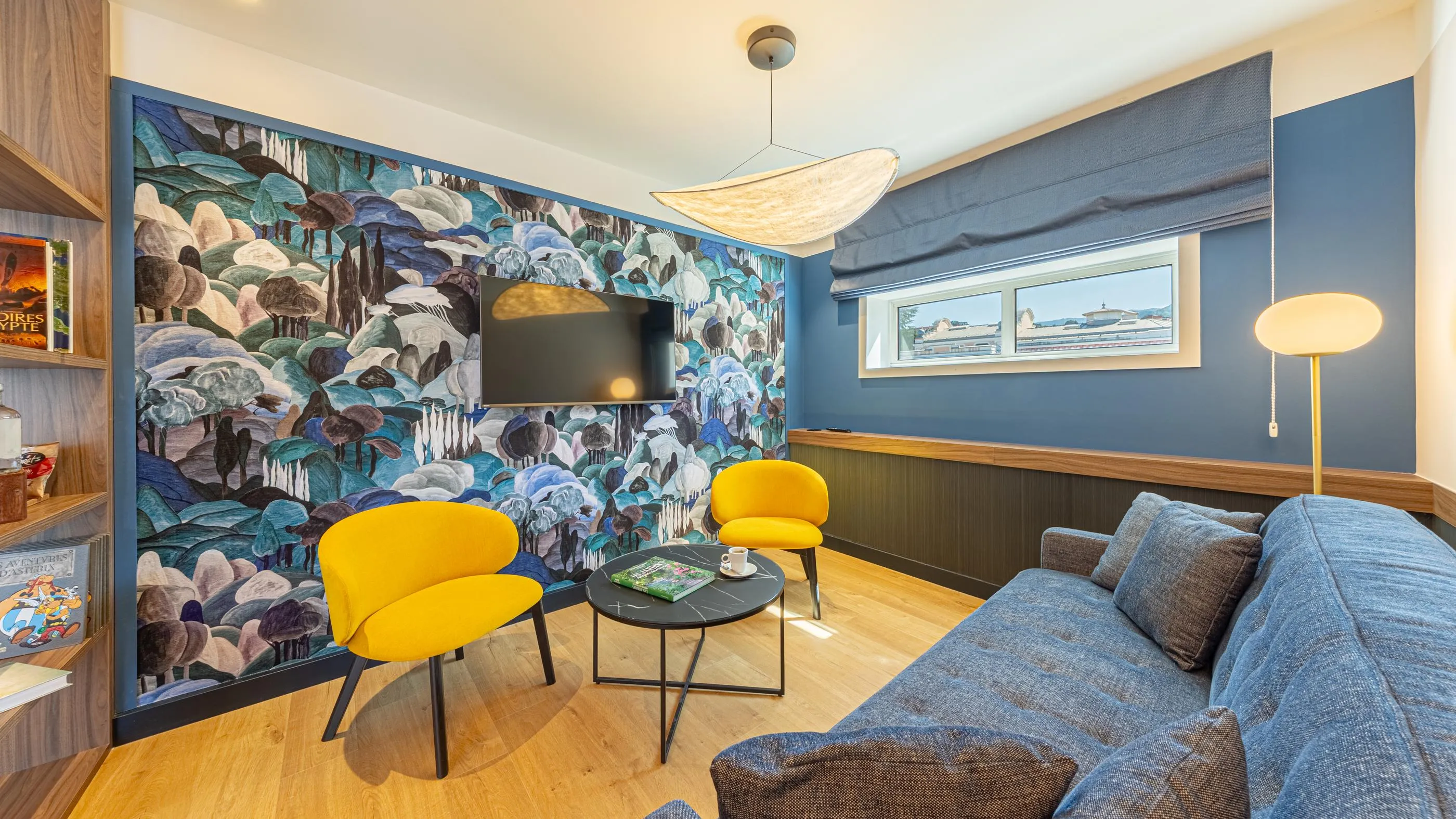 Salon avec canapé bleu, deux fauteuils jaunes, une table basse, un mur décoré d'un grand motif floral et une télévision