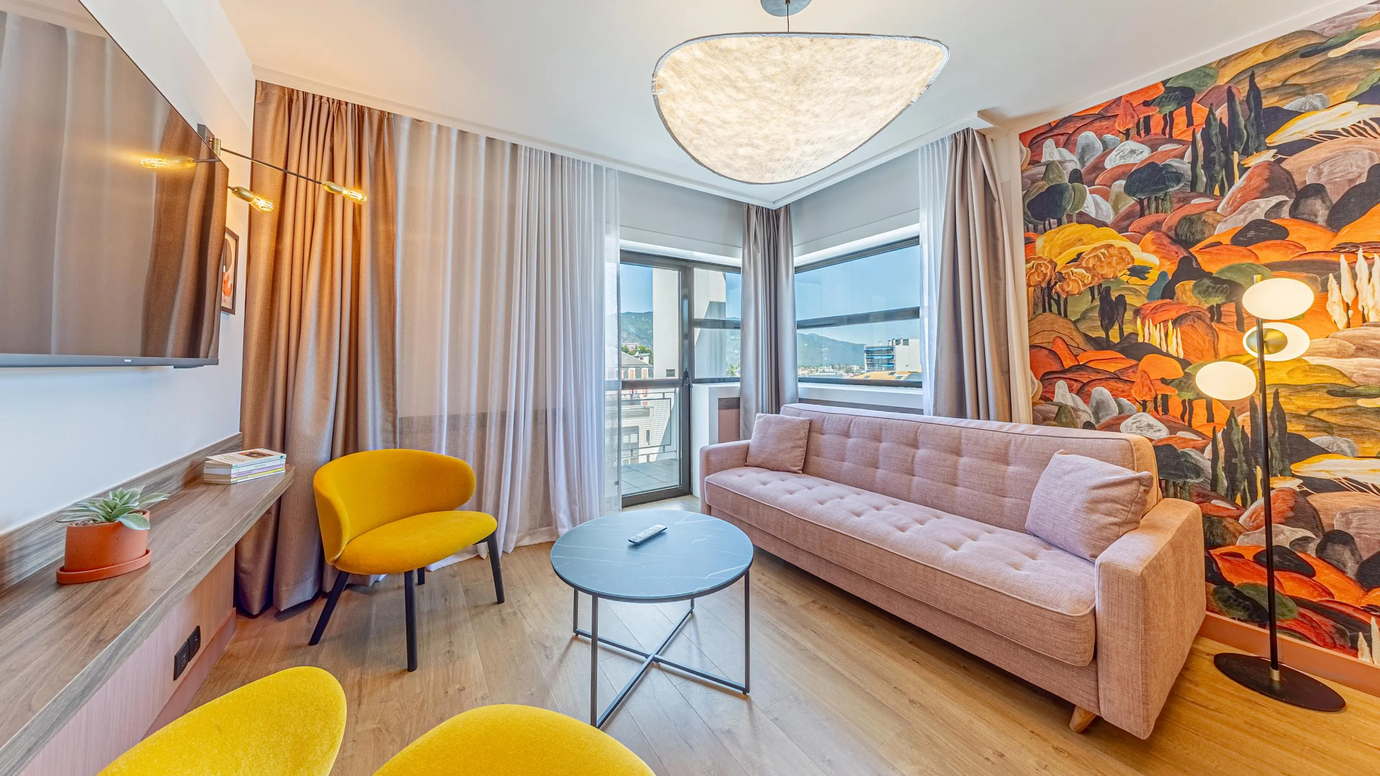 Salon moderne avec canapé beige, deux fauteuils jaunes, table basse, télévision et grande fenêtre avec vue sur la ville