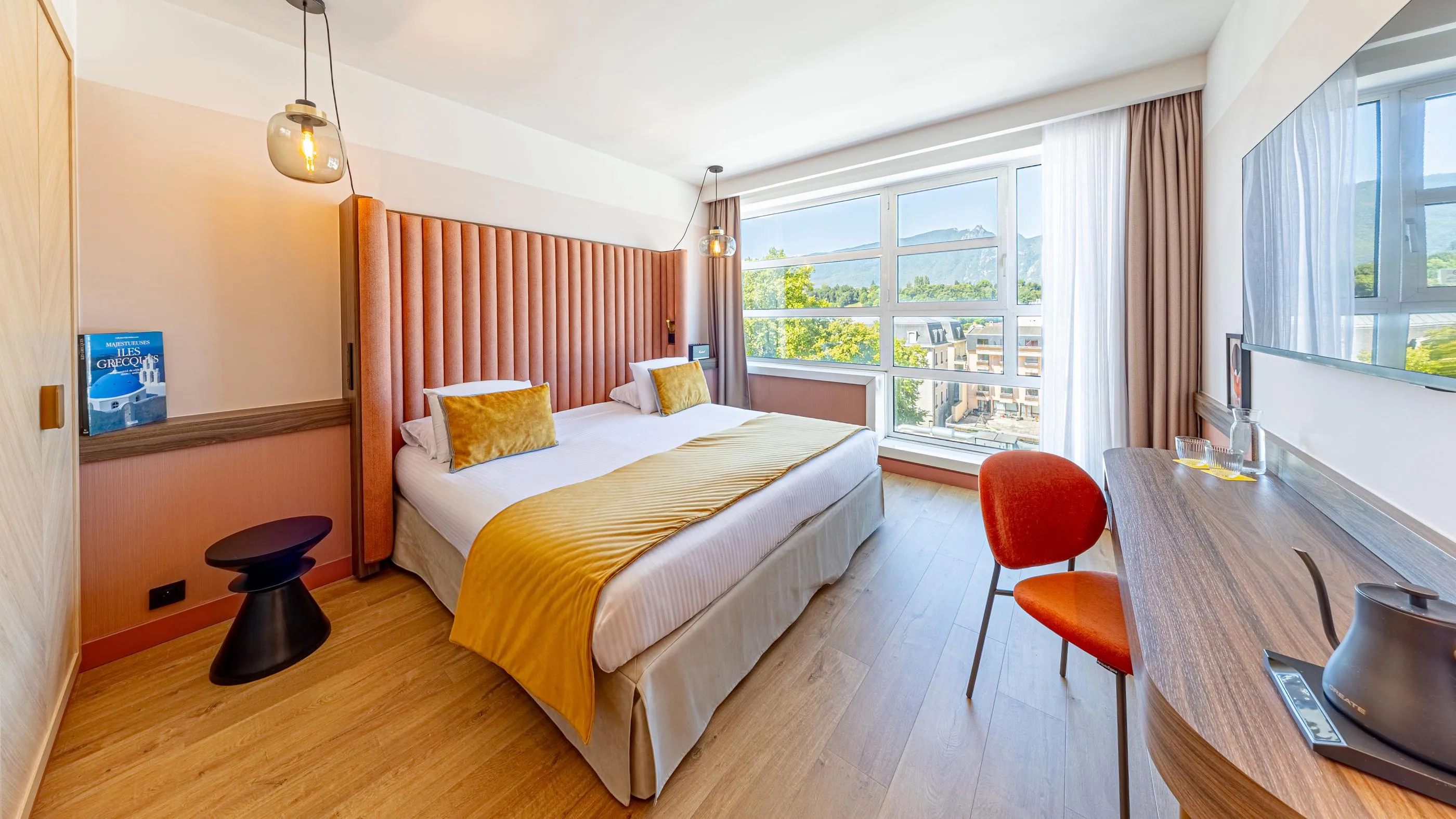 Chambre d'hôtel avec un lit double, une table de travail, une fenêtre offrant une vue sur des montagnes et des bâtiments, et des luminaires suspendus