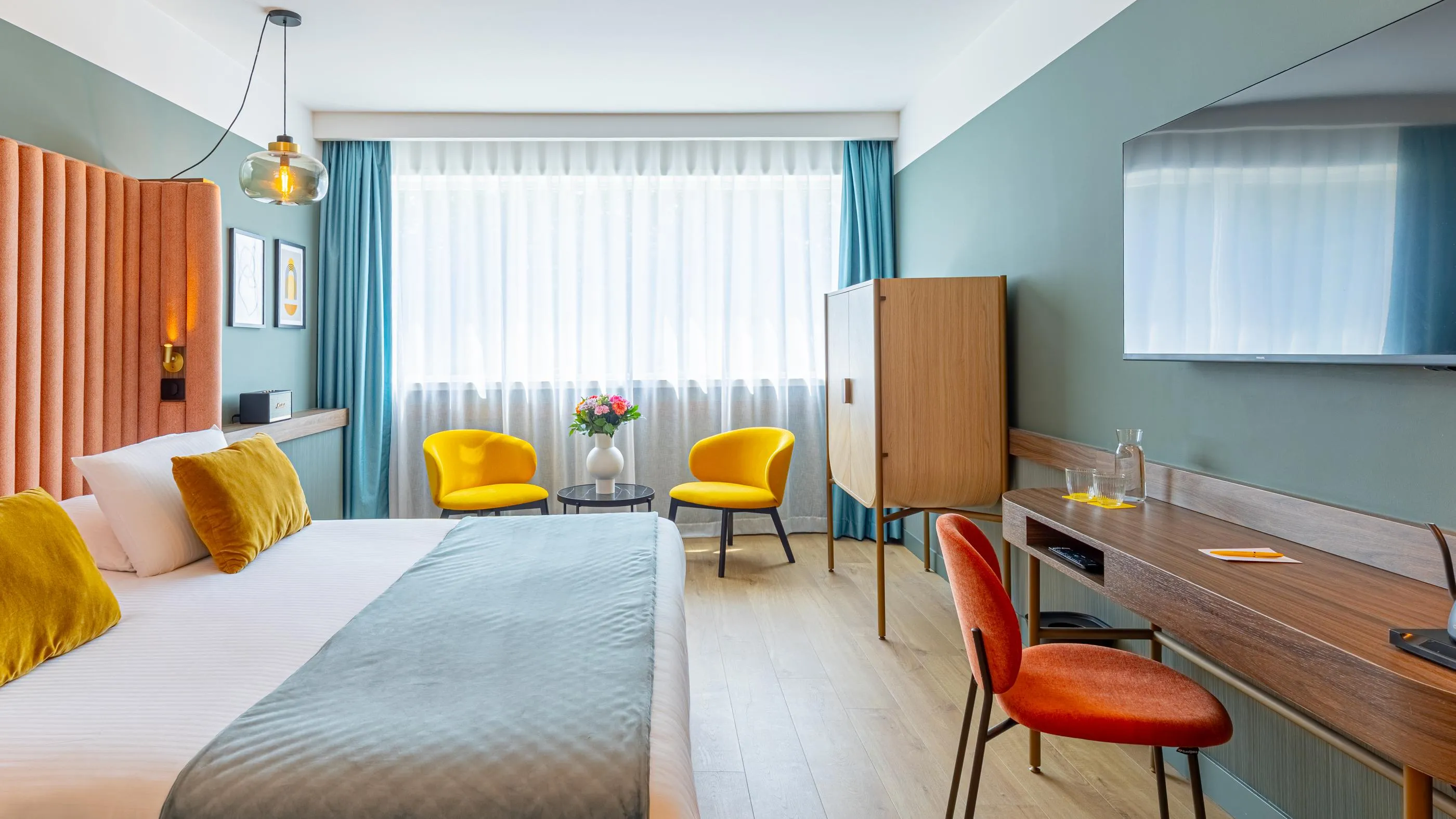 Chambre avec lit, deux chaises jaunes, bureau, télévision et grandes fenêtres avec rideaux