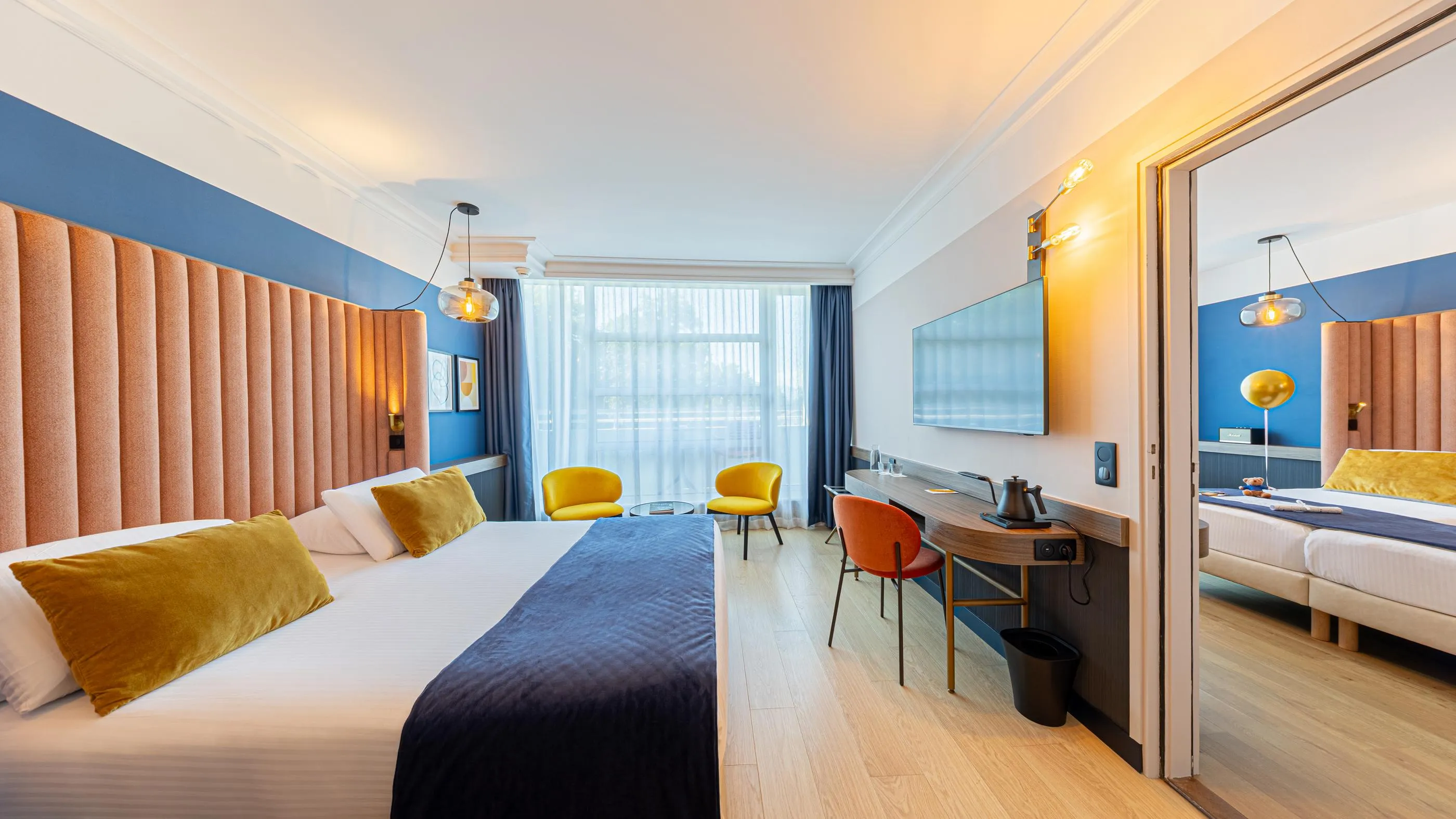 Chambre d'hôtel moderne avec deux lits, un coin salon et un bureau près d'une fenêtre avec vue sur un balcon