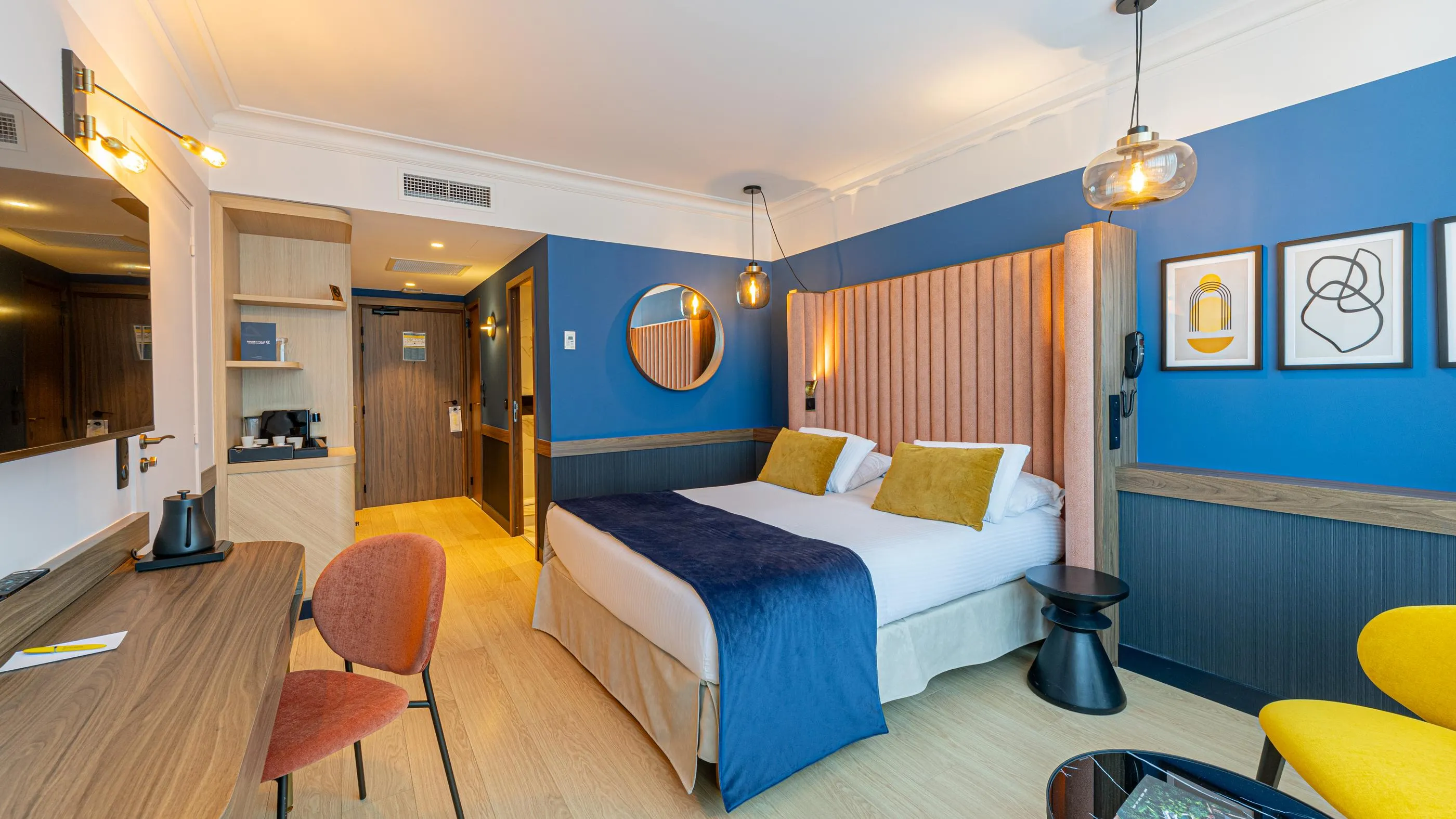 Chambre d'hôtel avec lit double, table de travail, deux chaises colorées et un coin salon, murs peints en bleu et décorations murales