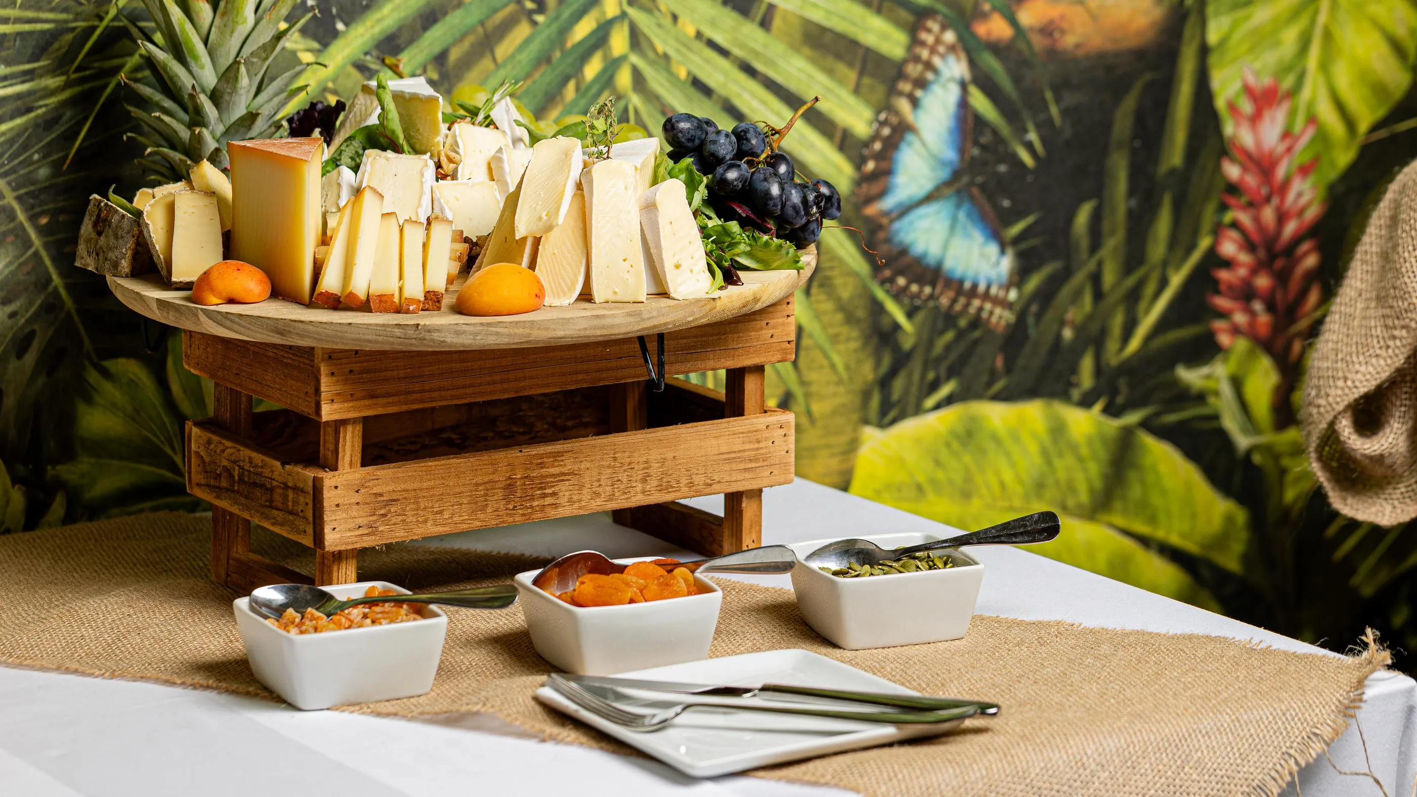 Un plateau de fromages et de fruits accompagné de trois petits bols de garnitures est posé sur une table recouverte d'une nappe en jute devant un fond de végétation tropicale