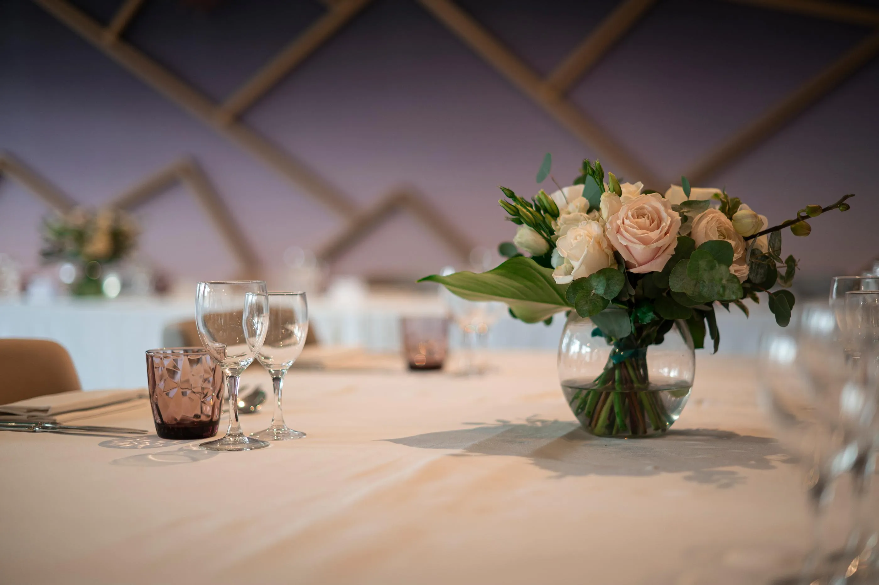 Table dressée avec des verres, une coupe en verre teinté et un vase contenant des roses pâles et des feuillages