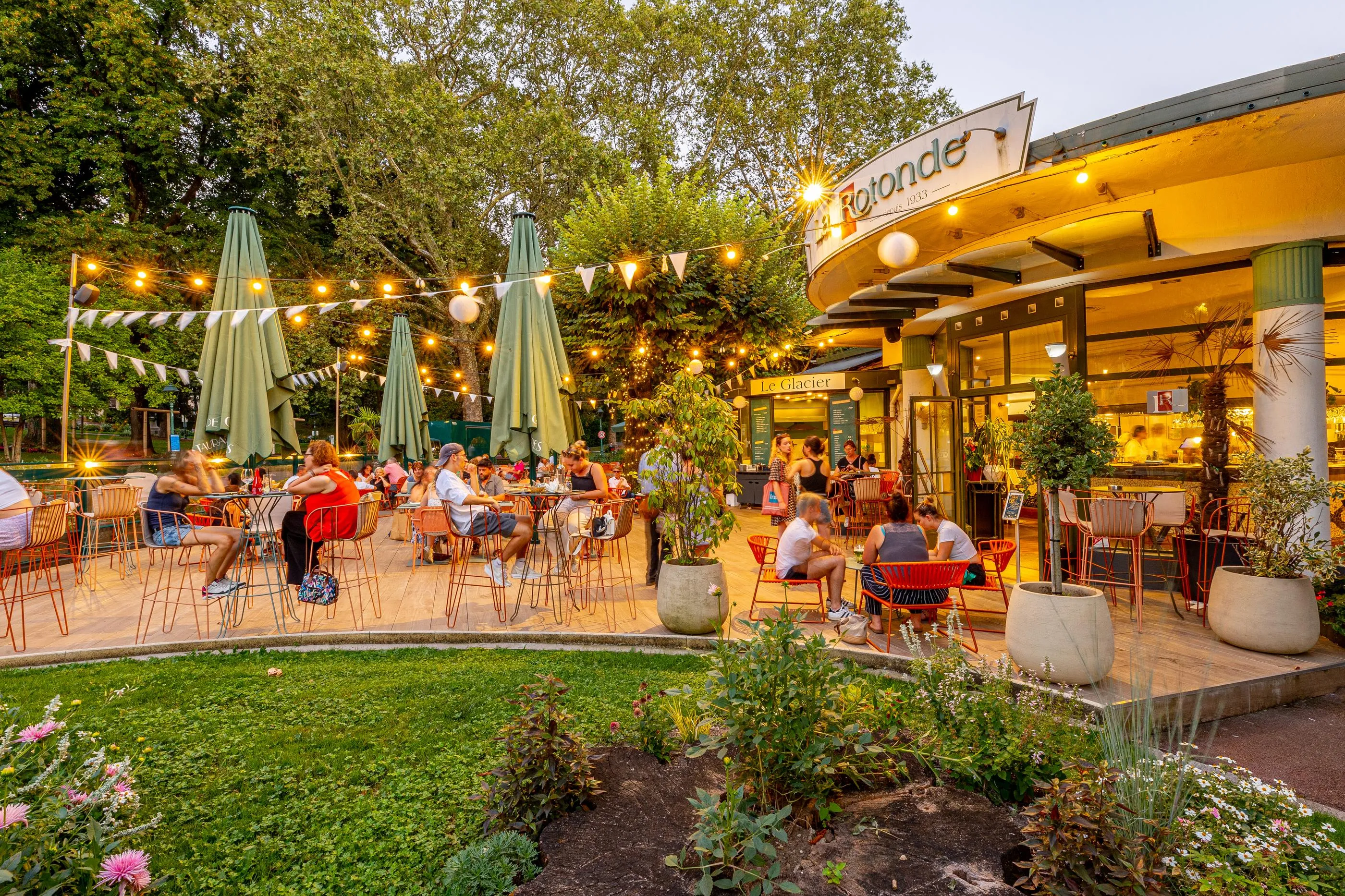 Terrasse de café en extérieur avec des clients assis à des tables sous des parasols, entourée de verdure et décorée de guirlandes lumineuses
