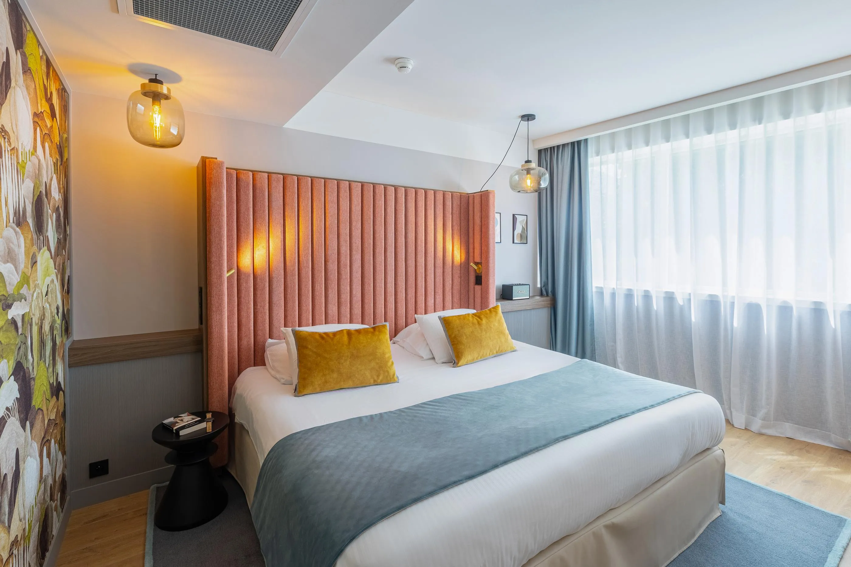 Chambre avec un lit double recouvert d'une couverture bleue et de coussins jaunes, des rideaux blancs et gris, des lampes murales et une décoration florale sur le mur