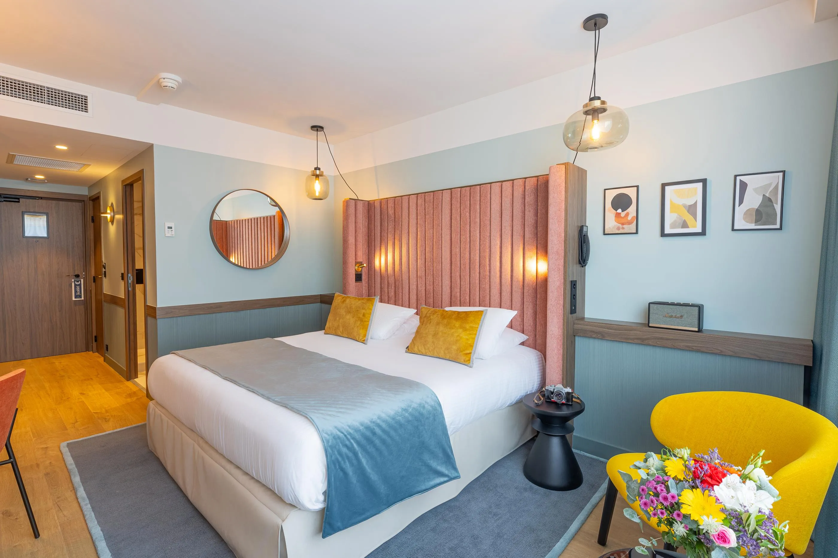 Chambre avec un lit double, une tête de lit en tissu, des coussins jaunes, une table de chevet, un fauteuil jaune et des fleurs sur la table