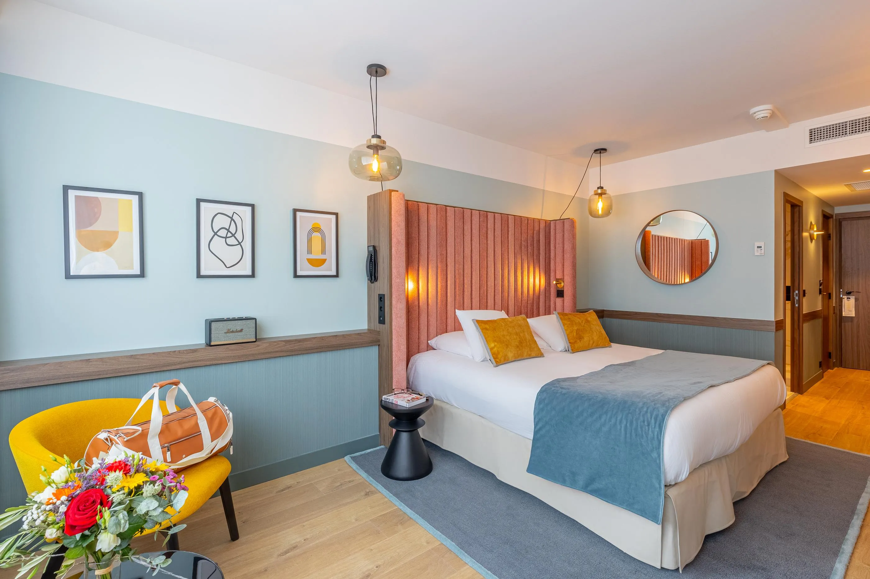Chambre avec un lit double, une tête de lit en tissu, des coussins jaunes, une couverture bleue, une table de chevet noire, un fauteuil jaune et des fleurs sur la table